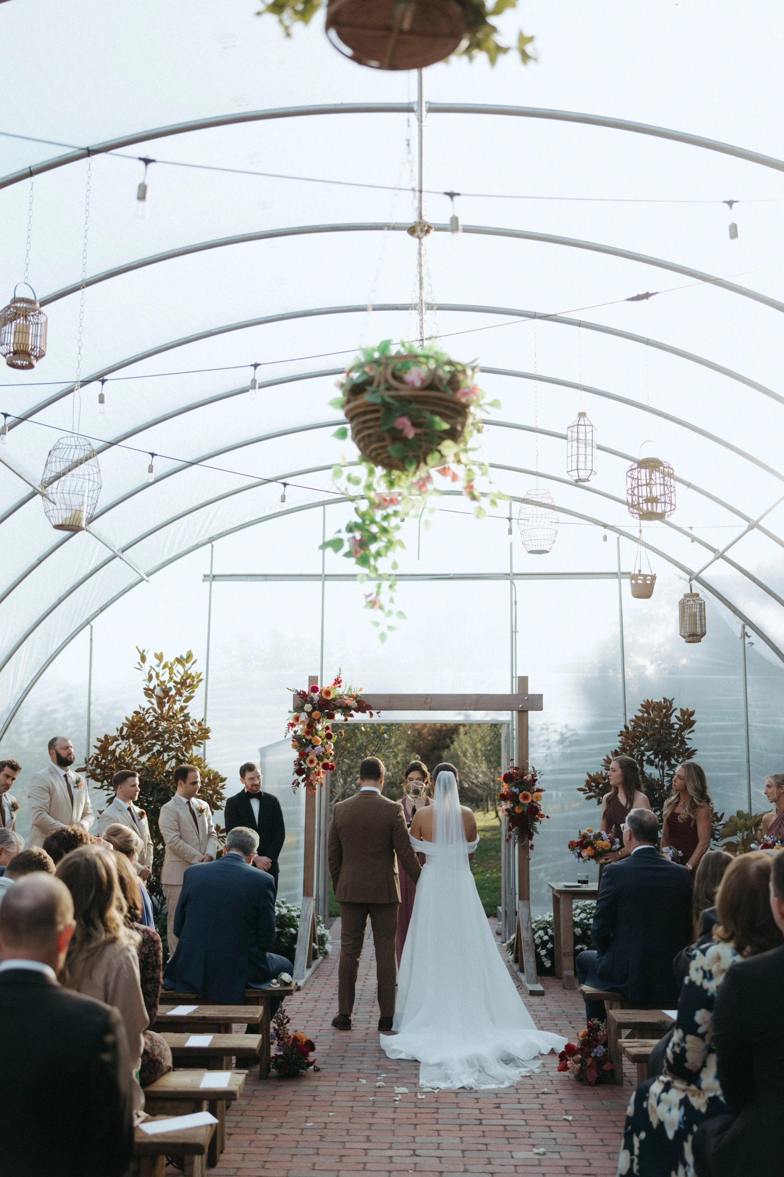 A wedding ceremony taking place inside a greenhouse with guests seated on benches, a bride and groom holding hands facing someone officiating, surrounded by floral arrangements and hanging lanterns.