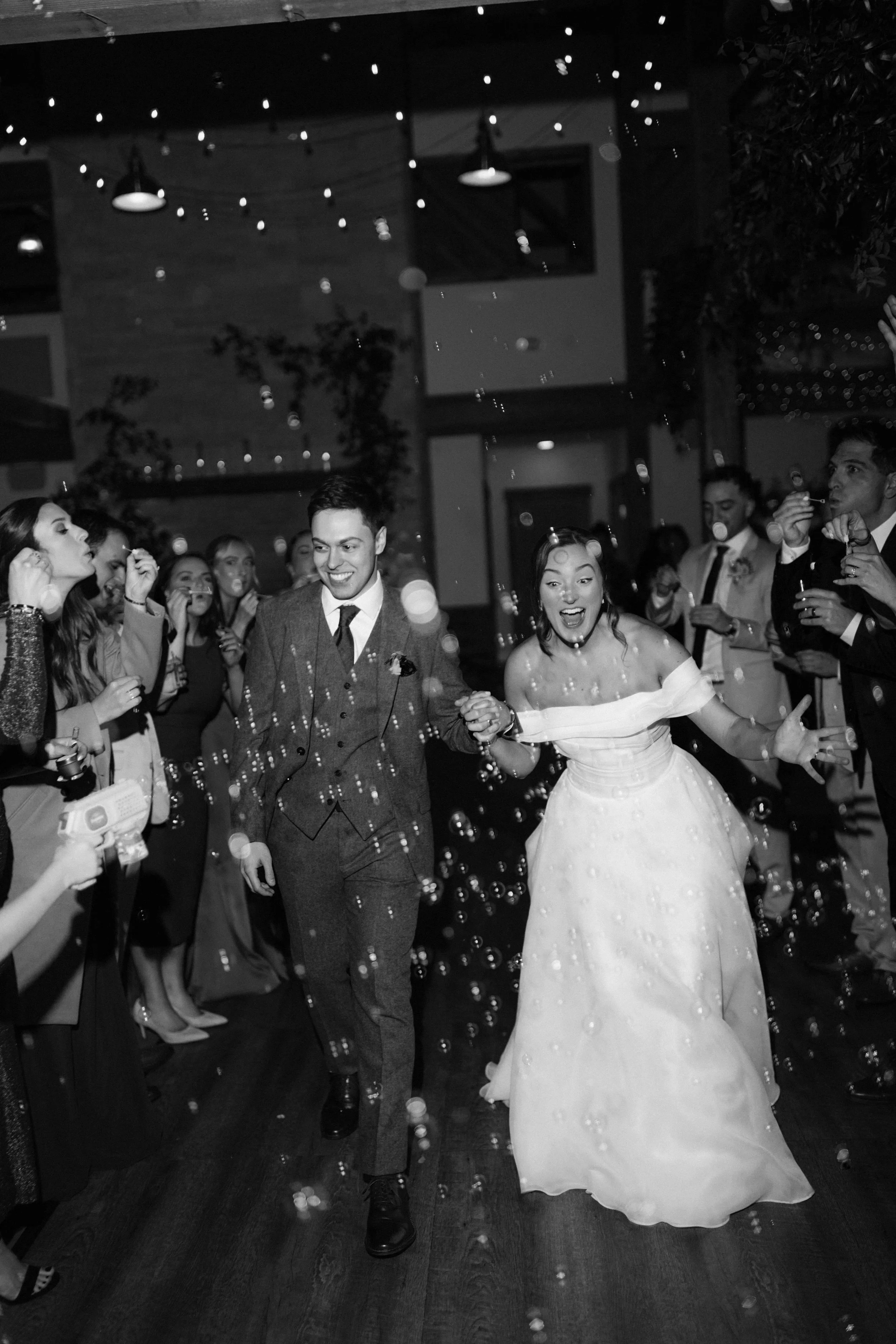 A bride and groom dancing at a wedding reception with guests surrounding them, in a decorated indoor venue with string lights and trees, and bubbles in the air.