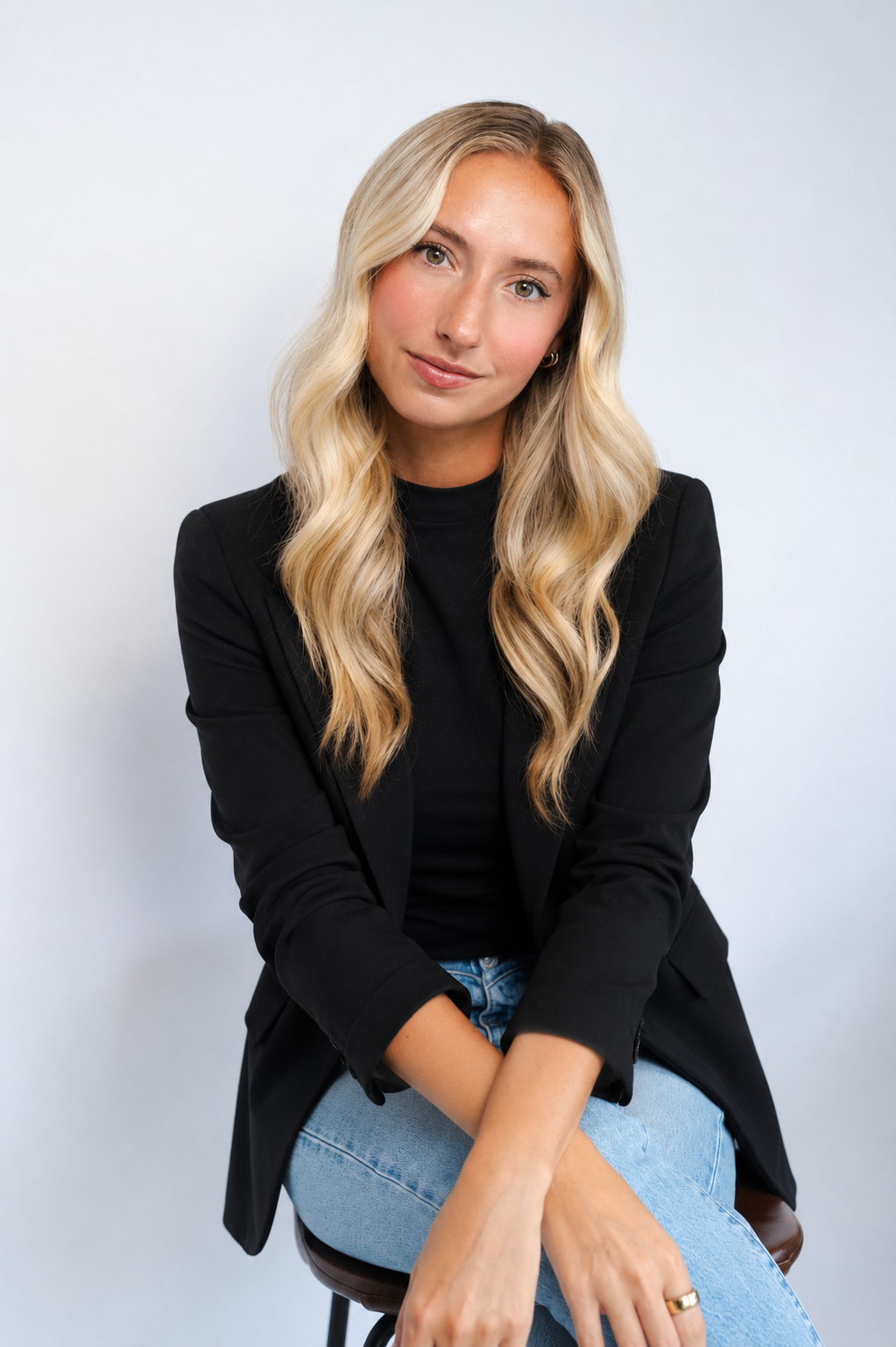 A young woman with long, wavy blonde hair, wearing a black blazer and light blue jeans, sitting on a brown stool against a plain white background.