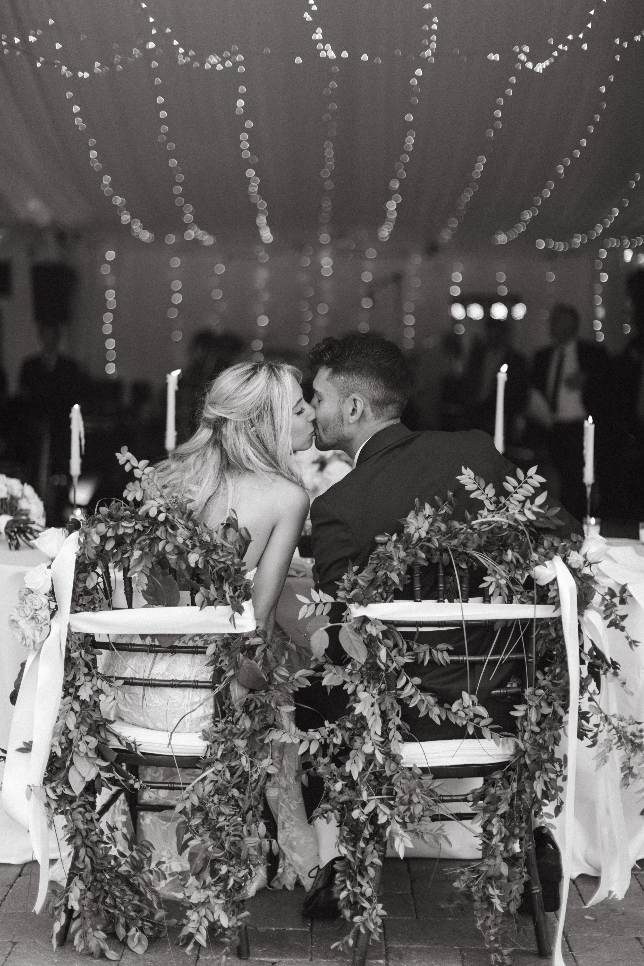 A black and white photo of a couple sharing a kiss, sitting at a decorated wedding table with greenery. The ceiling is adorned with string lights, and there are candles on the table.