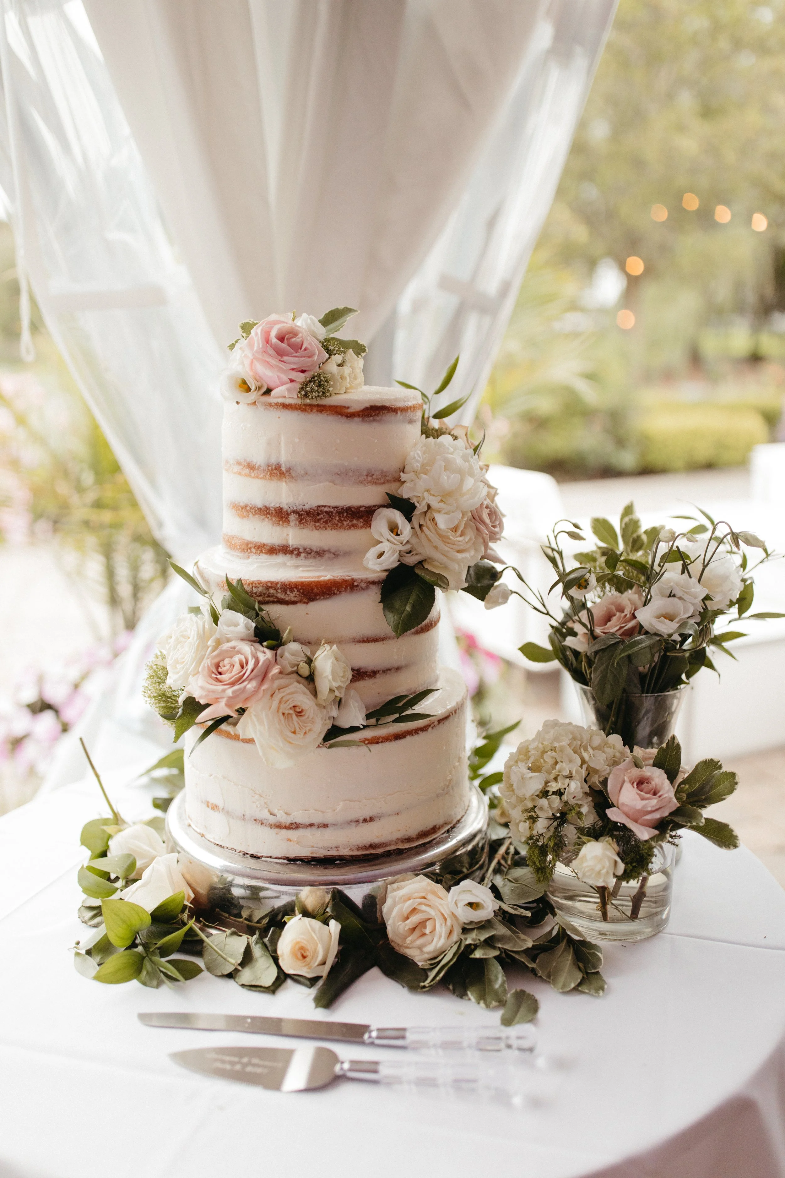 A tiered wedding cake decorated with fresh pink and white roses, surrounded by additional floral arrangements and greenery. The cake is on a glass stand with cake cutting utensils in front of it, set on a white table.