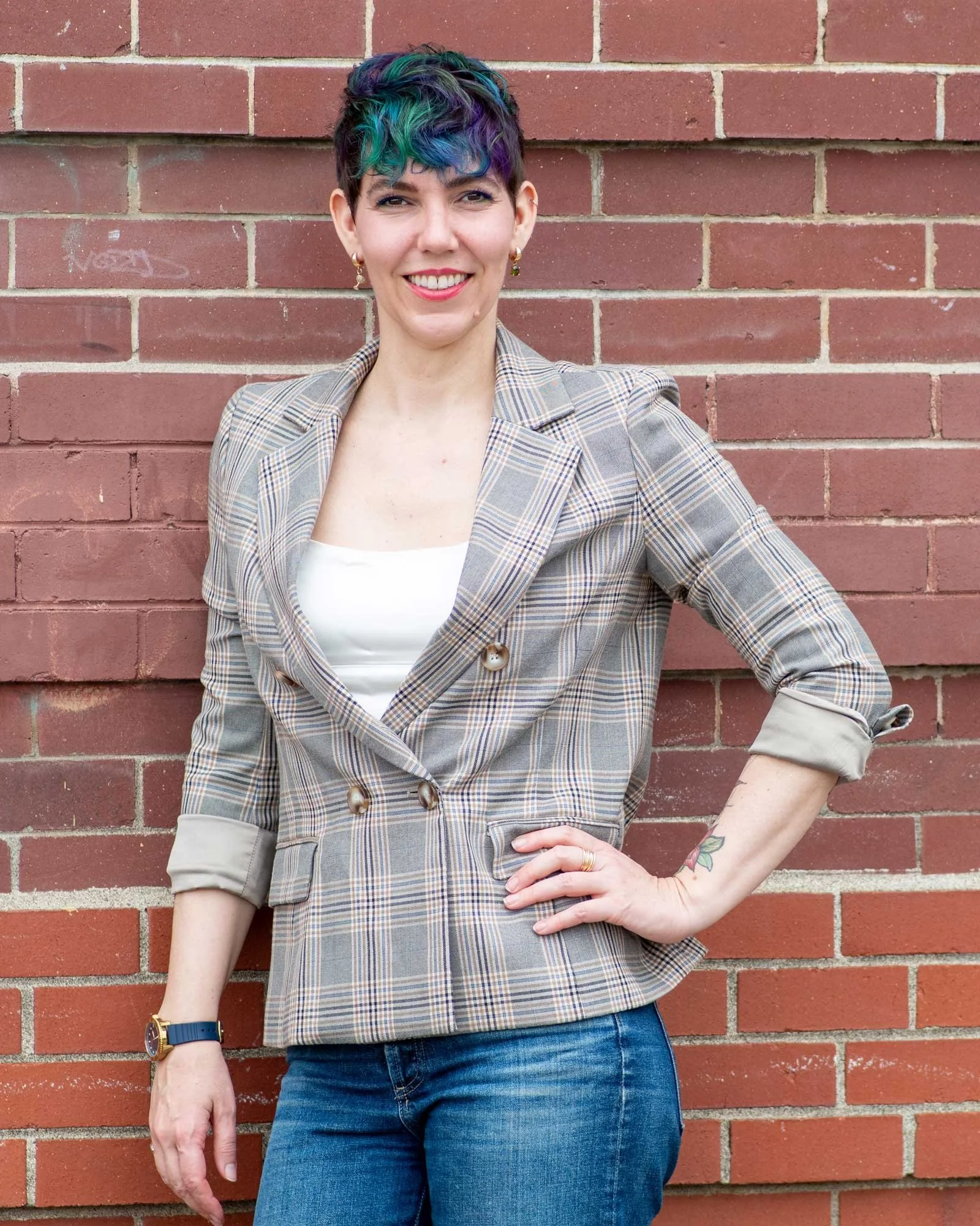 A woman with short, colorful hair standing in front of a brick wall, smiling and wearing a plaid blazer and blue jeans.
