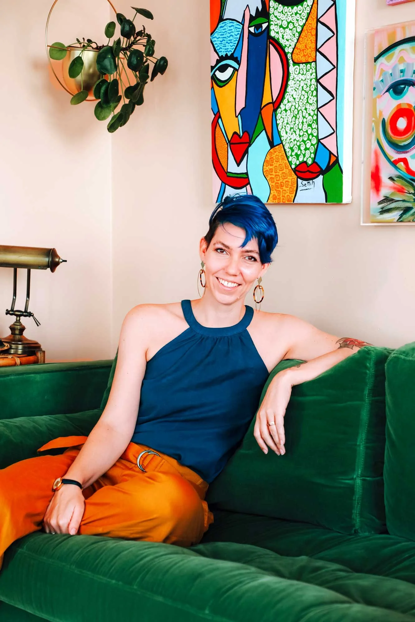 A woman with short blue hair and earrings sitting on a green velvet couch in a room decorated with colorful abstract art and a potted plant hanging on the wall.