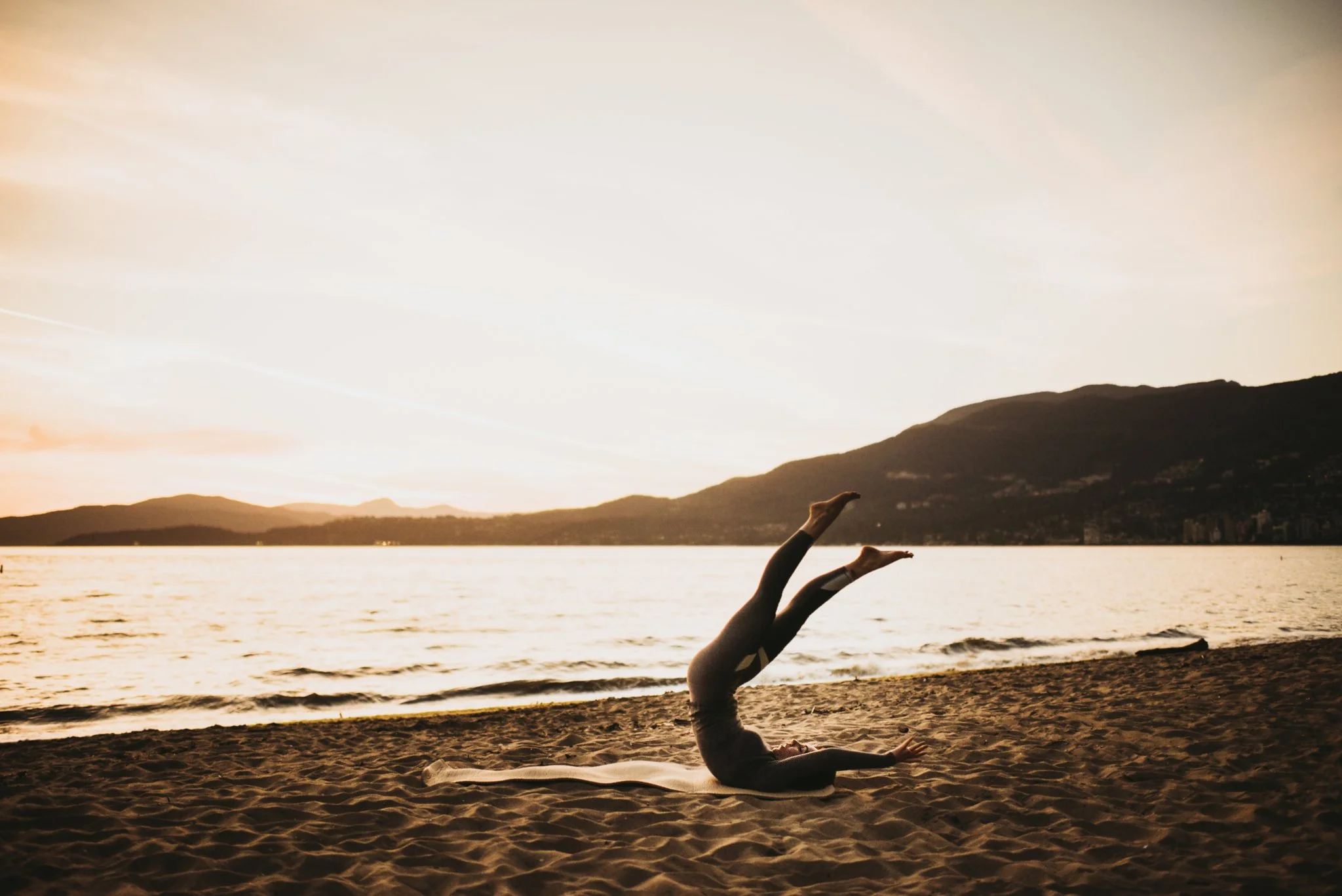 Person practicing yoga on the beach during sunset, with mountains and water in the background.