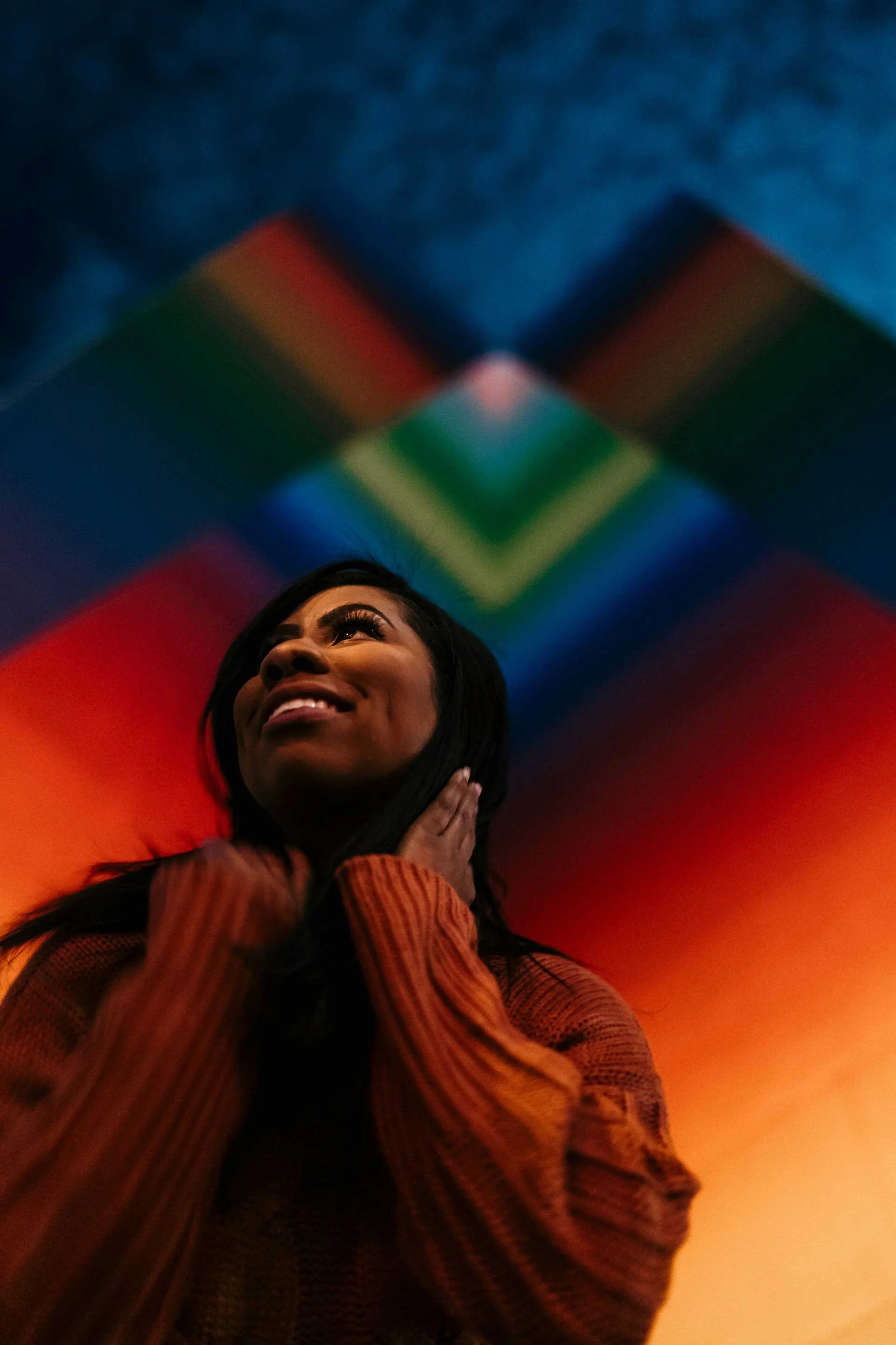 A woman with long dark hair, smiling, wearing a rust-colored, ribbed sweater, standing against a background with colorful, blurred lights in red, orange, green, blue, and purple.