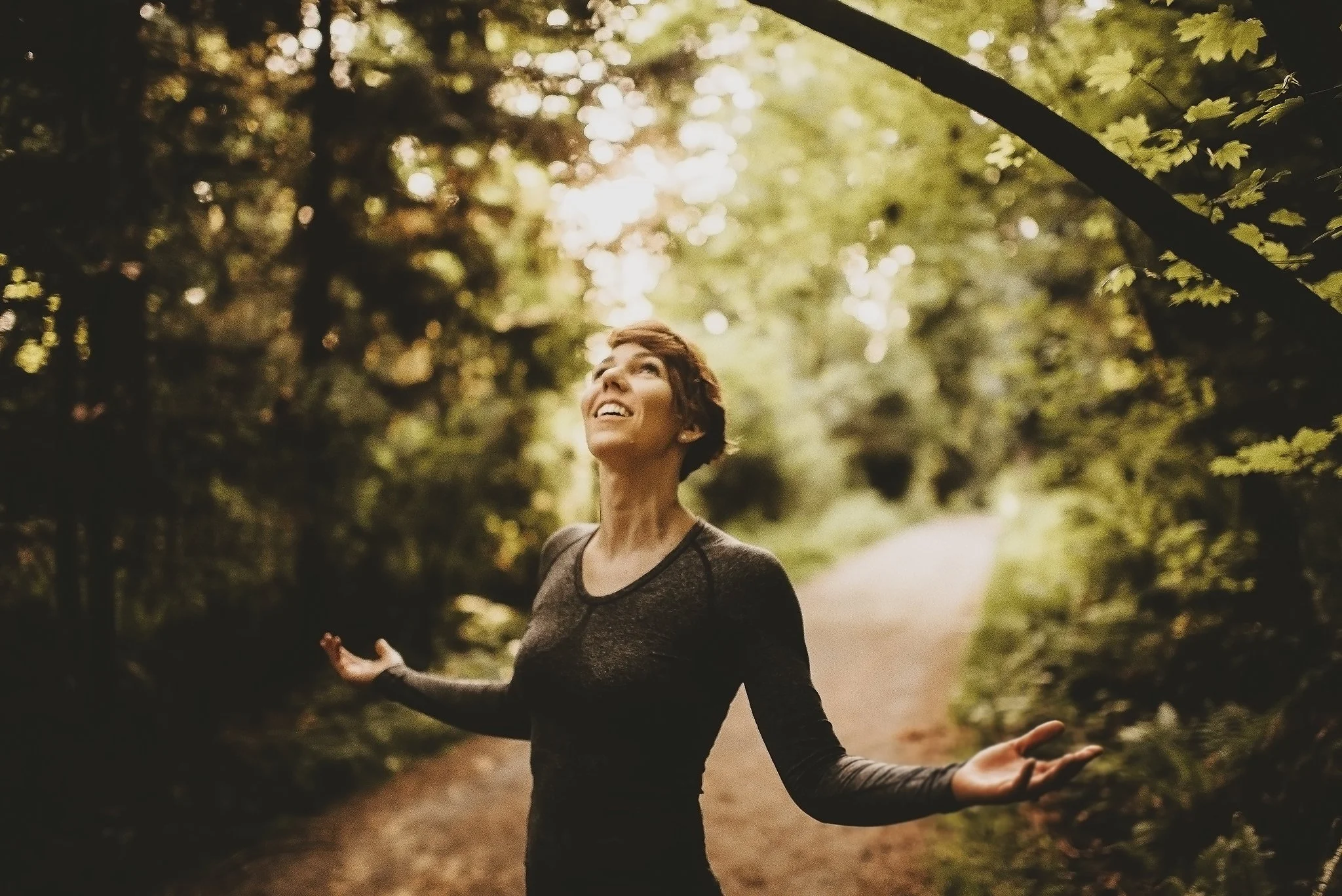 A woman in a black long-sleeve dress is standing outdoors on a path surrounded by trees with green leaves, smiling and looking up, with sunlight shining through the trees.