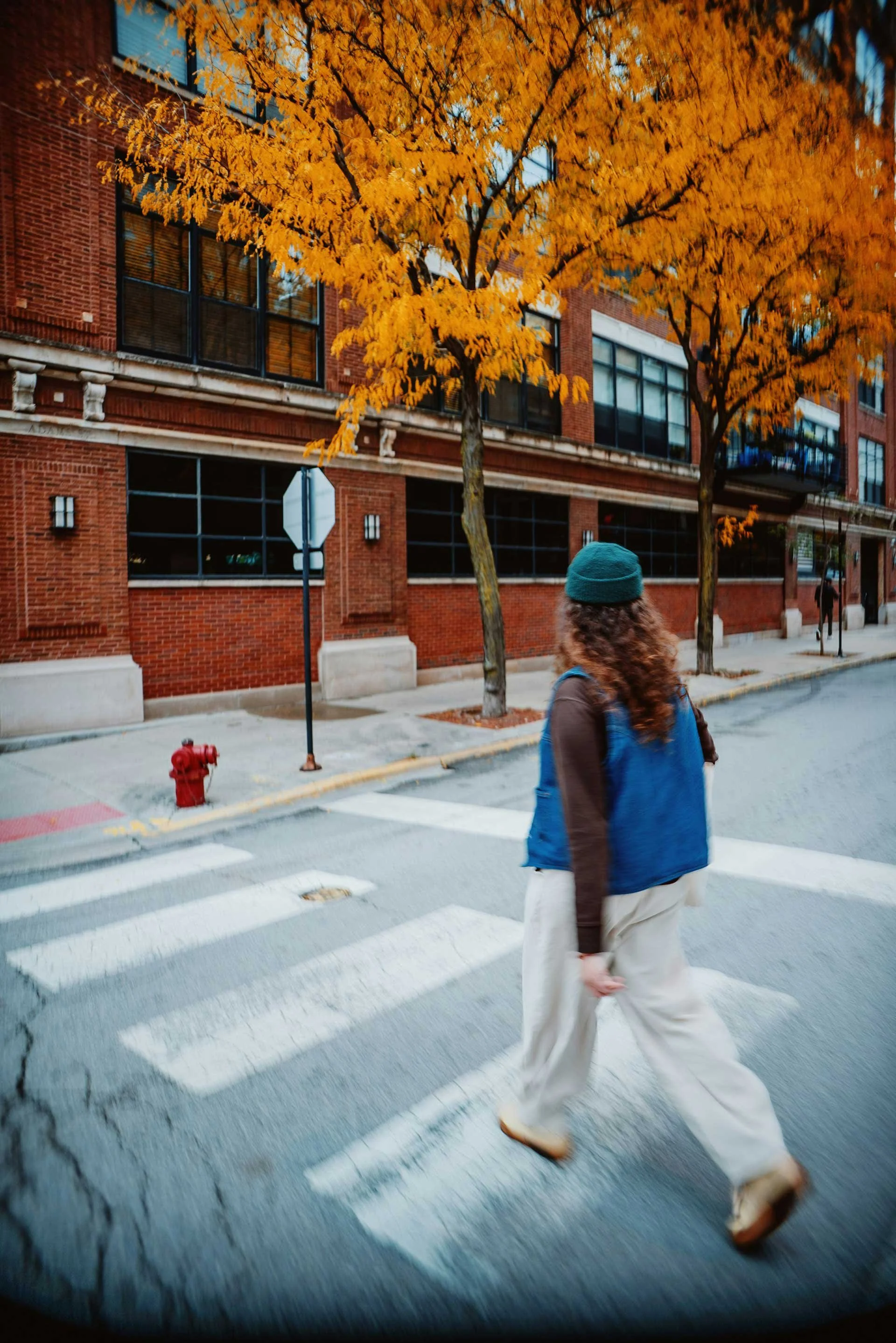 A person with curly hair, wearing a teal beanie, blue vest, and wide-leg pants crossing a city street on a pedestrian crosswalk during autumn. There are orange-leaved trees, a brick building with large windows, a stop sign, and a red fire hydrant.