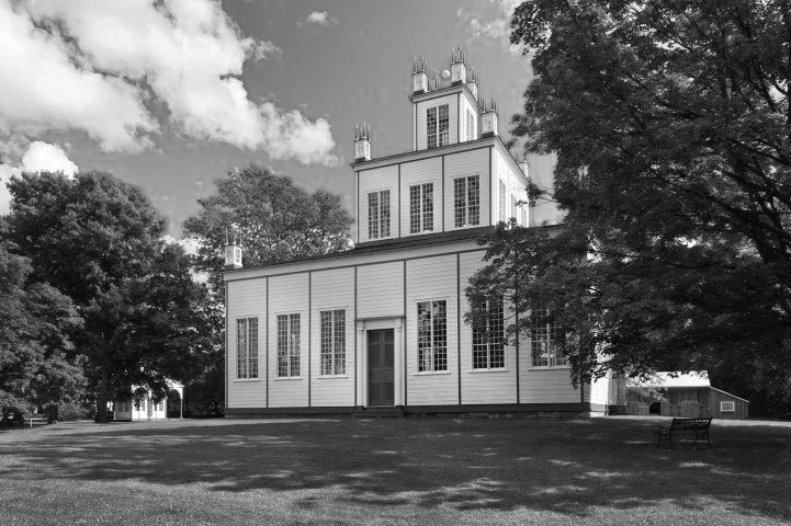 Historic wooden church with steeple, surrounded by trees and an open grassy area.