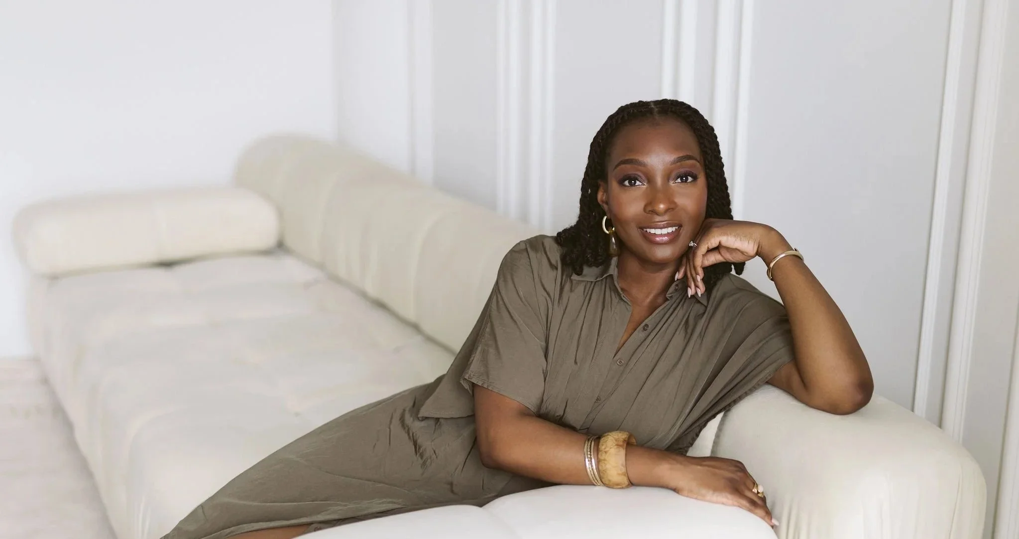 A woman with dark skin and braided hair is sitting on a white couch in a bright room, smiling at the camera. She is wearing a taupe-colored dress and gold jewelry.