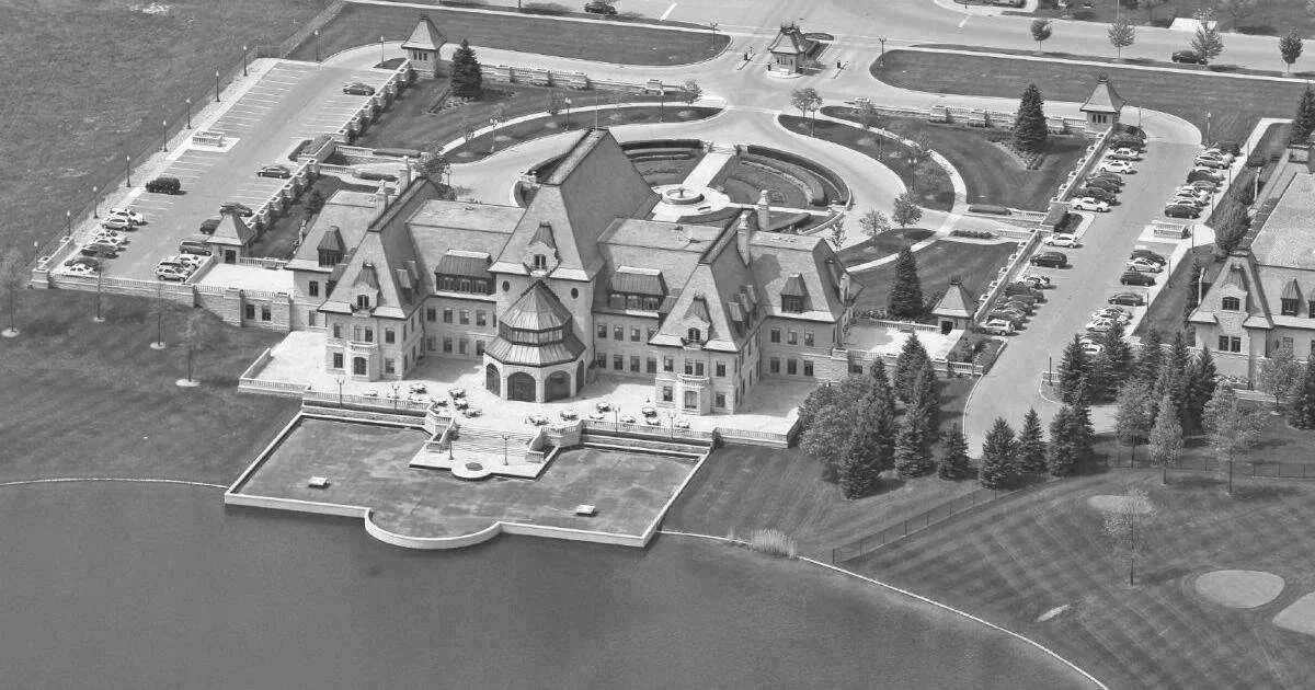 Aerial view of a large, castle-like mansion with multiple turrets, surrounded by a landscaped lawn, parking lots, and pathways, overlooking a body of water.