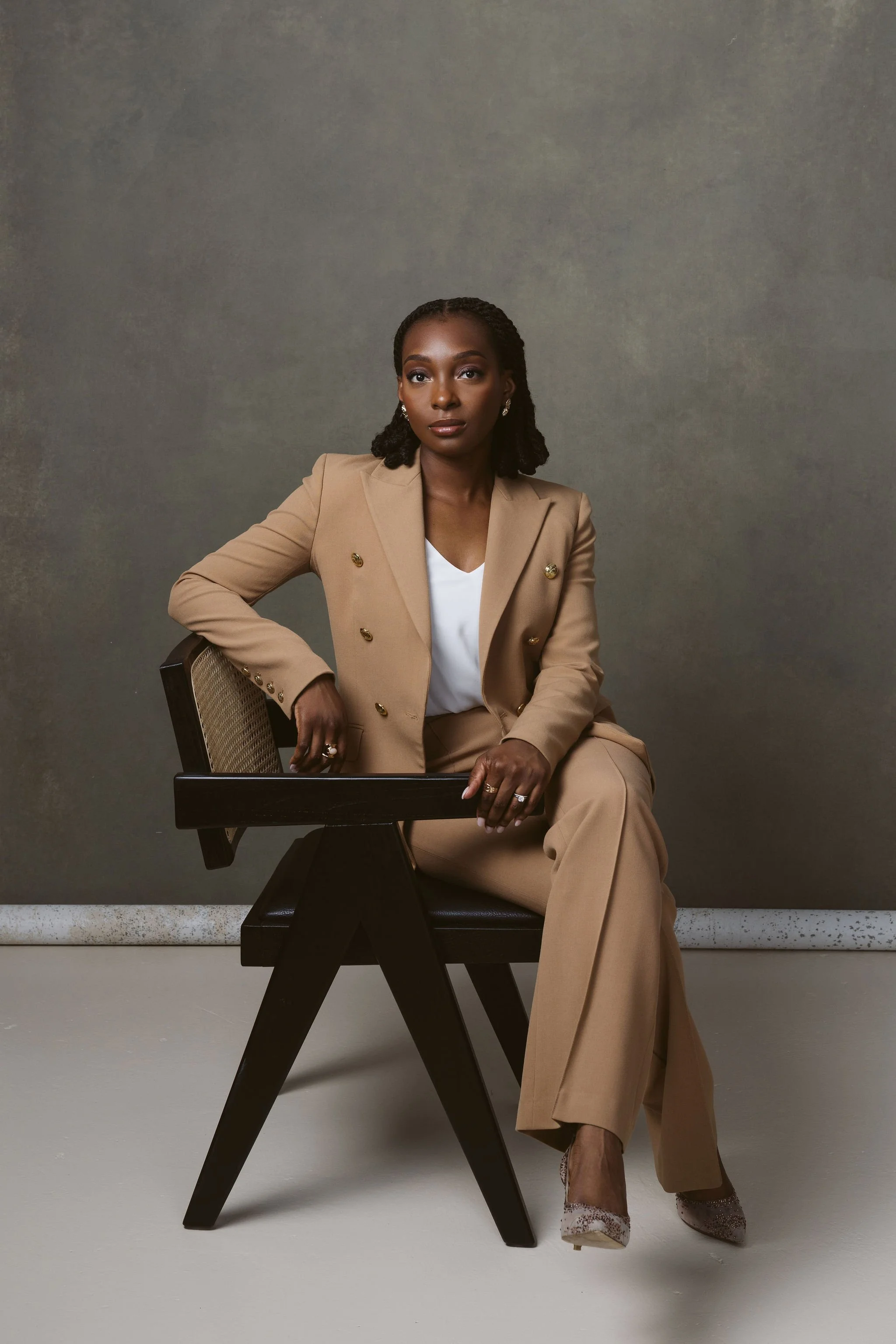 A woman with dark skin and braided hair sitting on a modern black wooden chair with a beige padded seat in front of a plain gray background. She is dressed in a beige blazer with gold buttons, matching beige pants, a white top, and sparkly high-heeled shoes. She has earrings and a ring on her right hand.