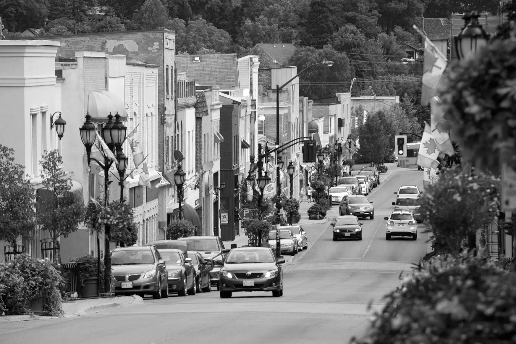 A black and white photo of a small town street with parked cars, buildings, hanging flower baskets, and trees in the background.