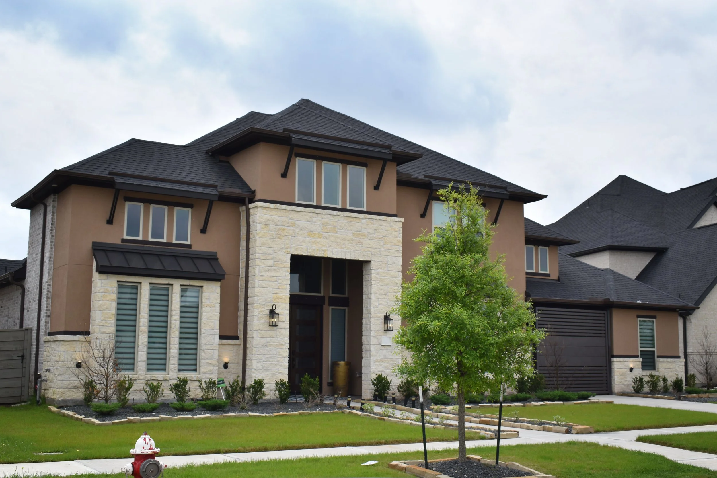 Front view of a modern multi-story house with stone and stucco exterior, large windows, and a dark shingle roof, with a landscaped lawn and young trees.