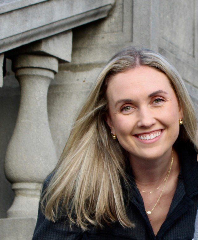 A woman with long blonde hair smiling, wearing gold jewelry, standing next to stone balustrades.