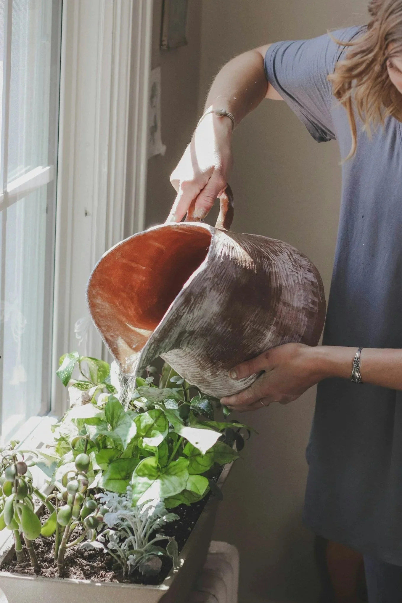 Woman pouring water from a large seashell-shaped planter filled with green houseplants, near a window.