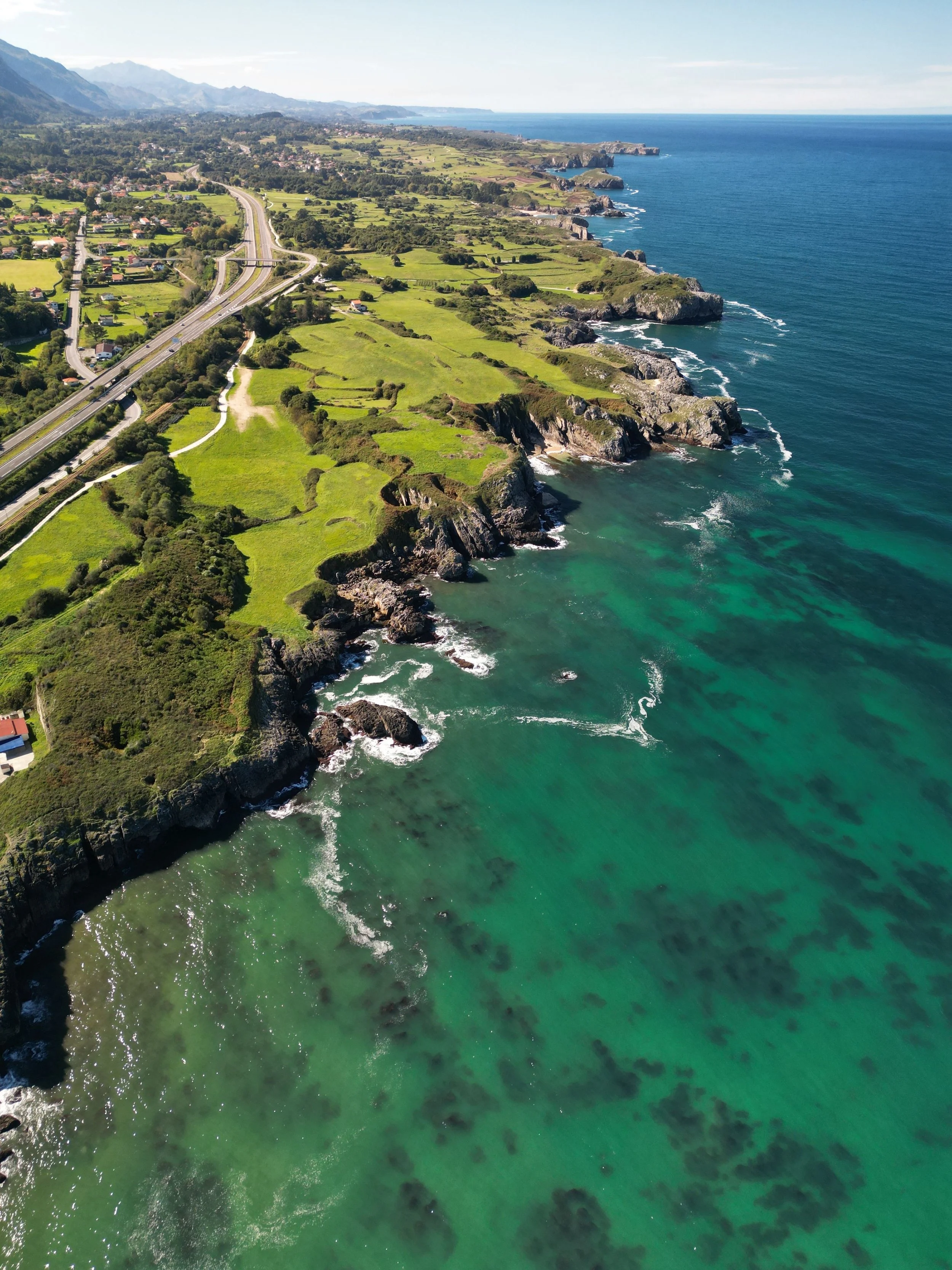 Aerial view of a scenic coastal highway curving along lush green cliffs and rocky turquoise Atlantic shorelines in Spain.