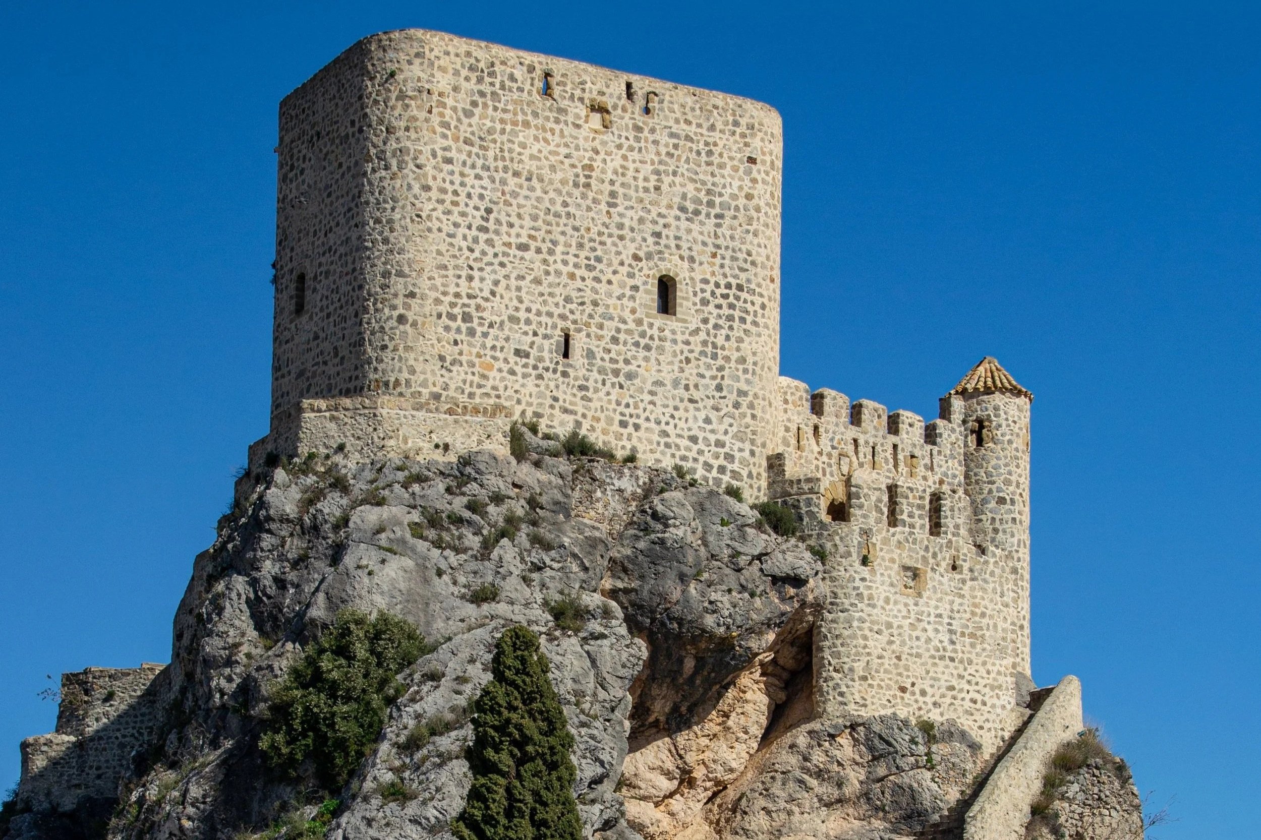 Looking up at a majestic stone medieval castle perched high atop a rocky hill under a clear blue sky.