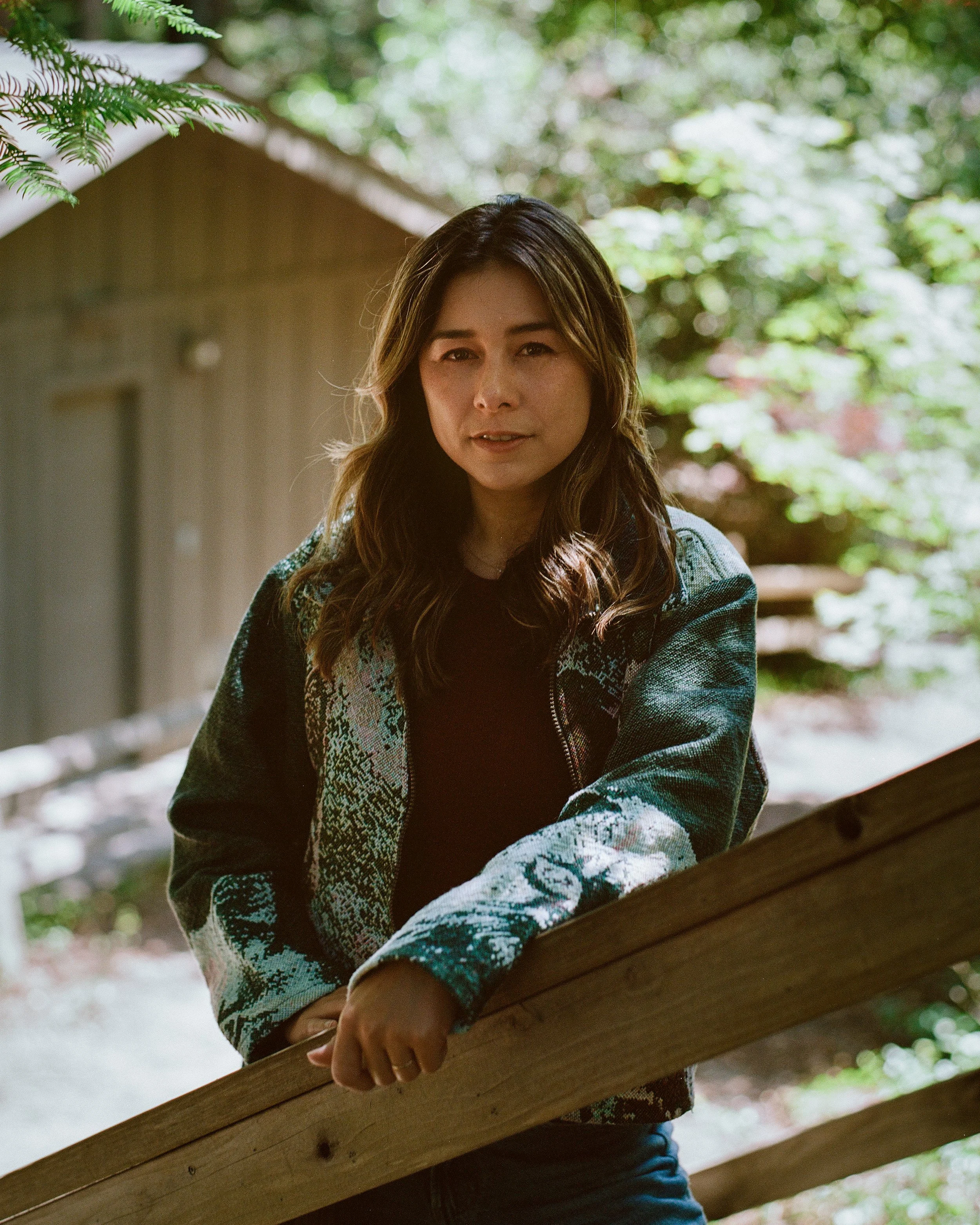 A woman with long, wavy brown hair and light skin leaning on a wooden railing outdoors, surrounded by greenery and trees in the background. A writer and author based in Los Angeles California. Romantasy, fantasy, and romance books.