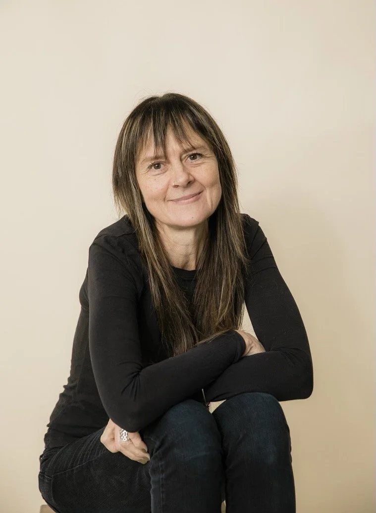 Woman with brown hair and a black long-sleeve shirt sitting against a plain beige wall.