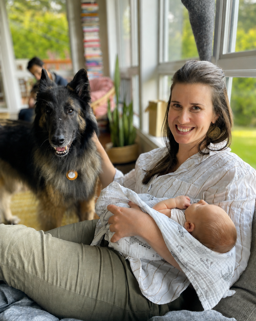A woman with a baby in her arms sitting on a couch, smiling at the camera, with a black and tan dog standing next to her indoors near a large window.