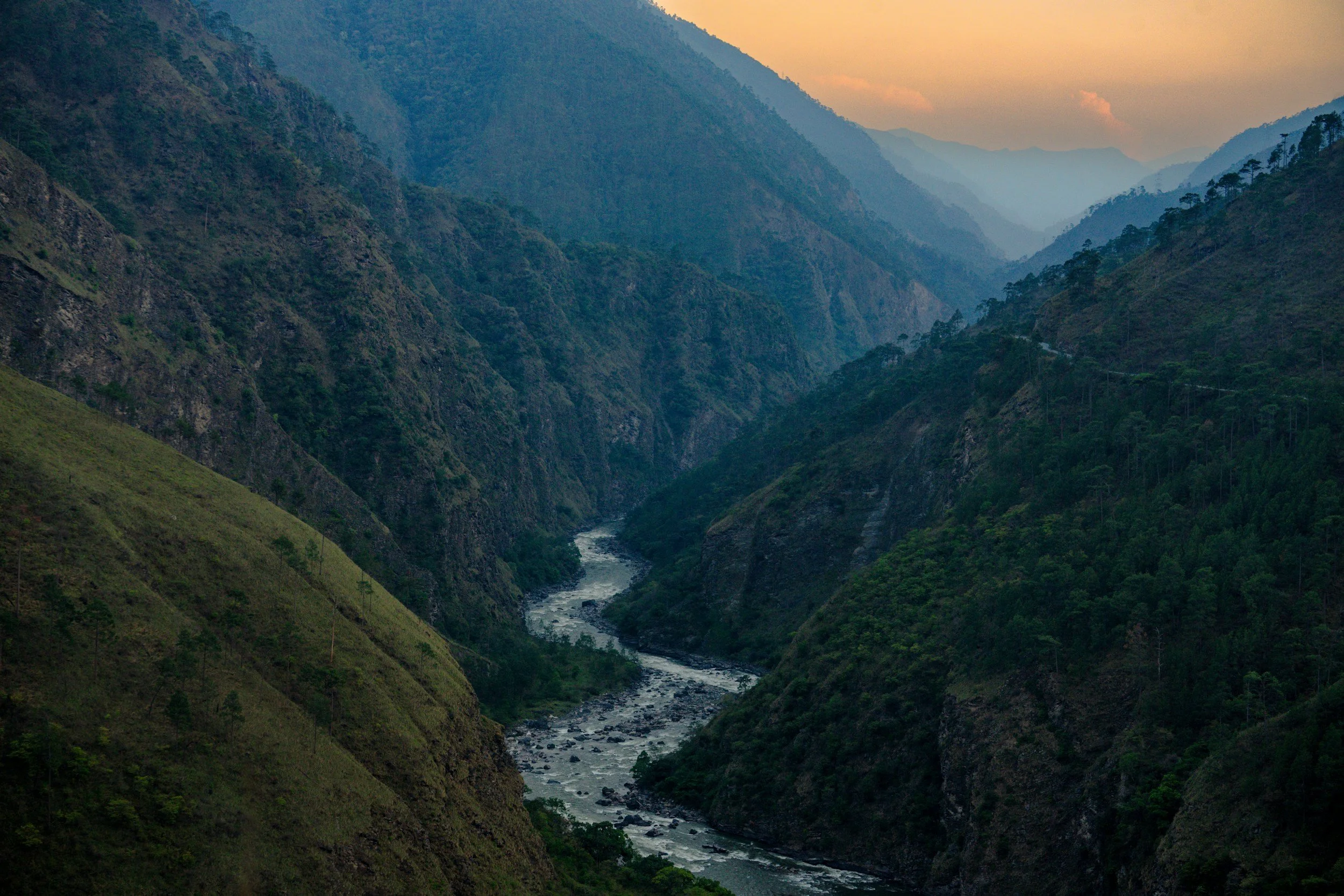 A scenic view of a deep river valley surrounded by tall, green mountains under a hazy sky at sunset.