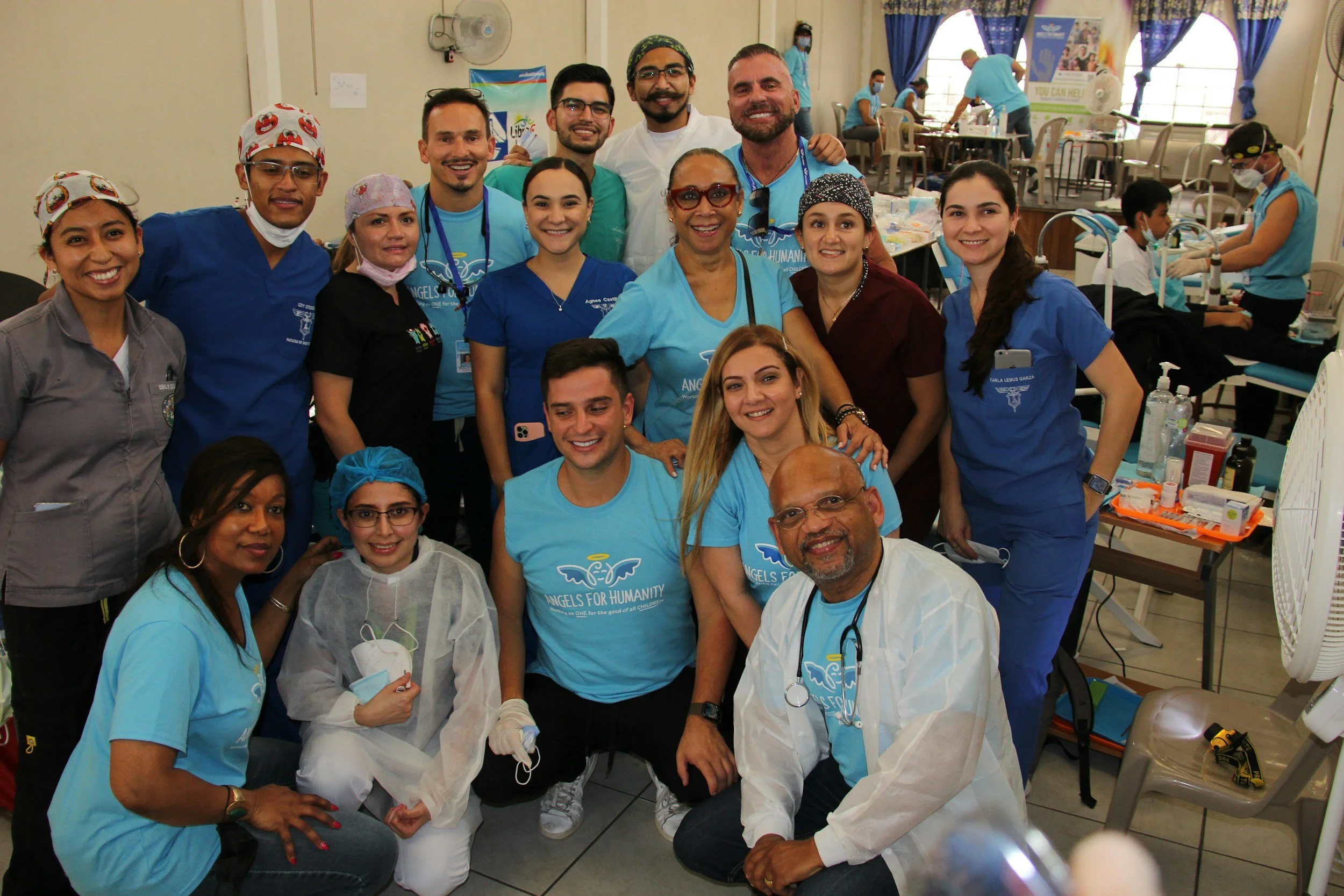 Group of healthcare workers and volunteers in a medical setting. They are smiling and posing for the photo, some wearing scrubs and medical masks. The background shows a clinical environment with medical equipment and other healthcare staff working.