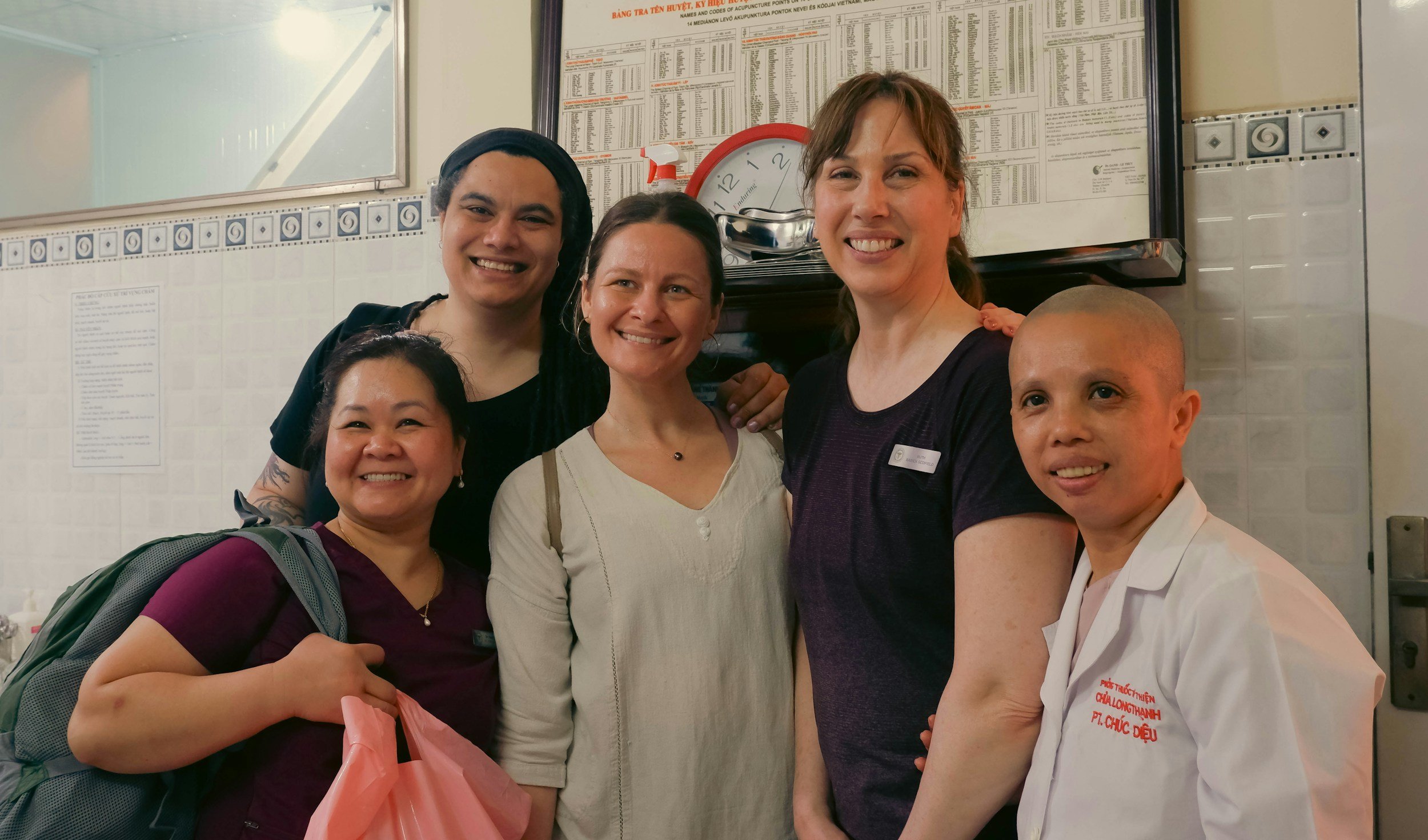 Group of five women and one man posing together inside a building, smiling at the camera, with a wall-mounted clock and a large chart or list in the background.