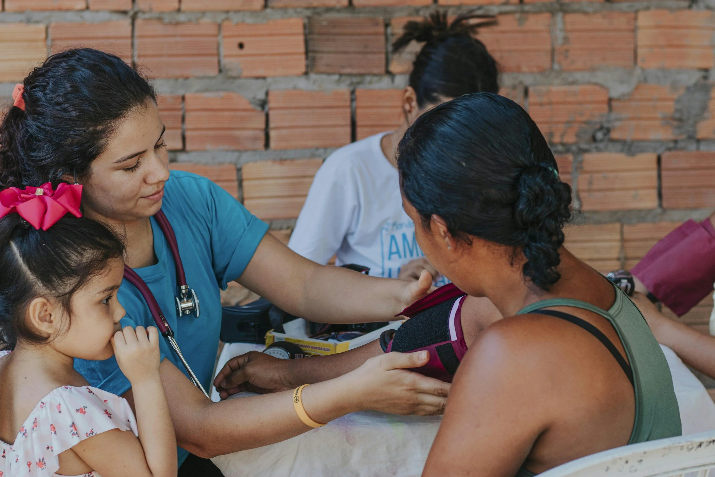 A healthcare worker, wearing a blue uniform and with a red hair bow, is assisting a woman who is receiving a medical check-up. The woman is seated and has dark hair pulled back, and there is a young girl with a pink hair bow observing the scene. The background features a brick wall.