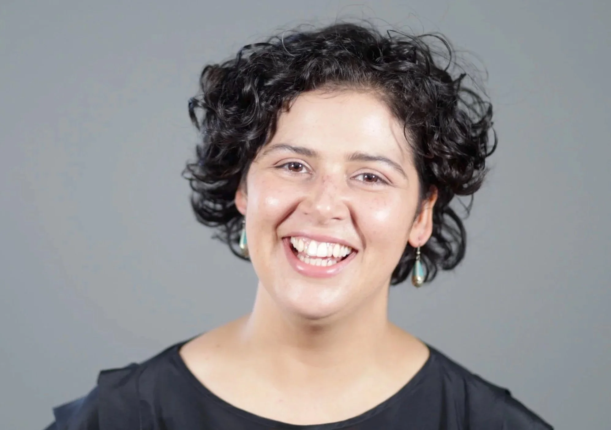 Close-up of a smiling woman with short curly dark hair, wearing earrings, against a plain gray background.