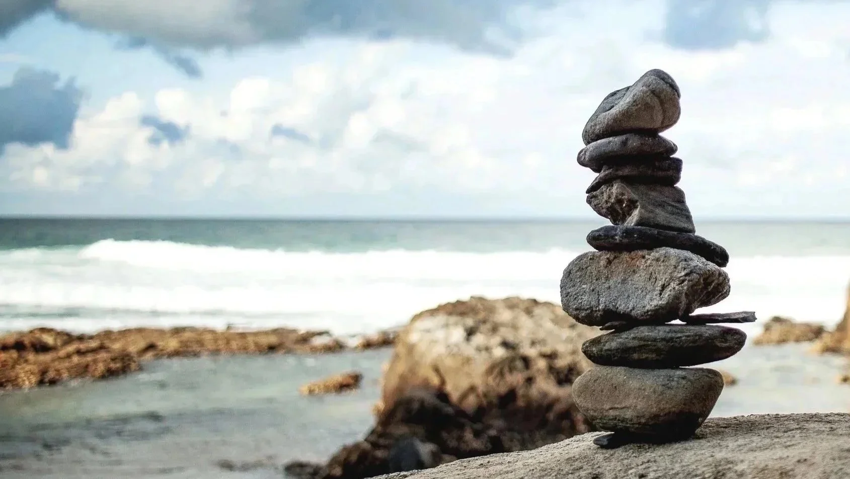 A stack of nine stones balanced on a rock on a beach with ocean waves and cloudy sky in the background.