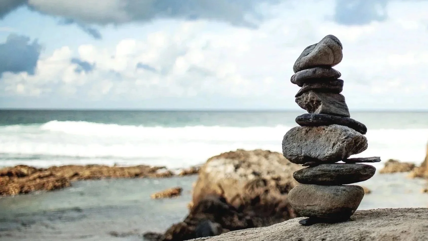 A stack of flat stones balanced on a larger rock on a beach, with ocean waves and cloudy sky in the background.
