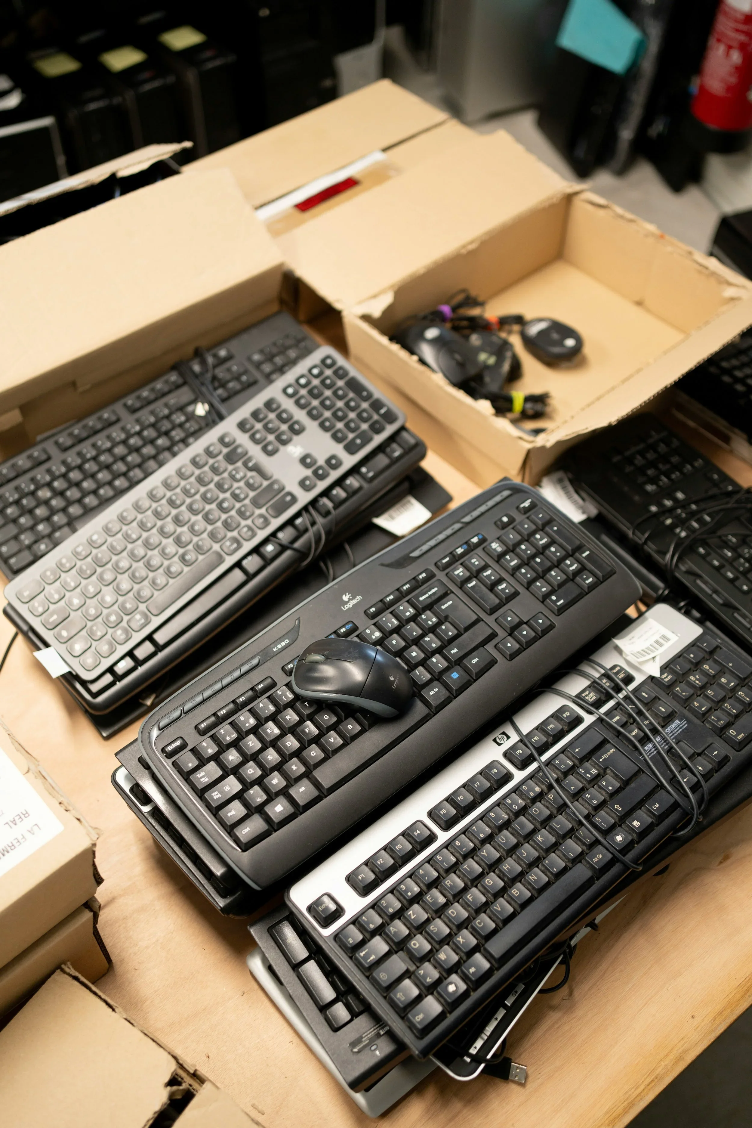 A table with multiple computer keyboards and a mouse, with a cardboard box containing car keys and key fobs. In the background, there are shelves with black boxes and a fire extinguisher.
