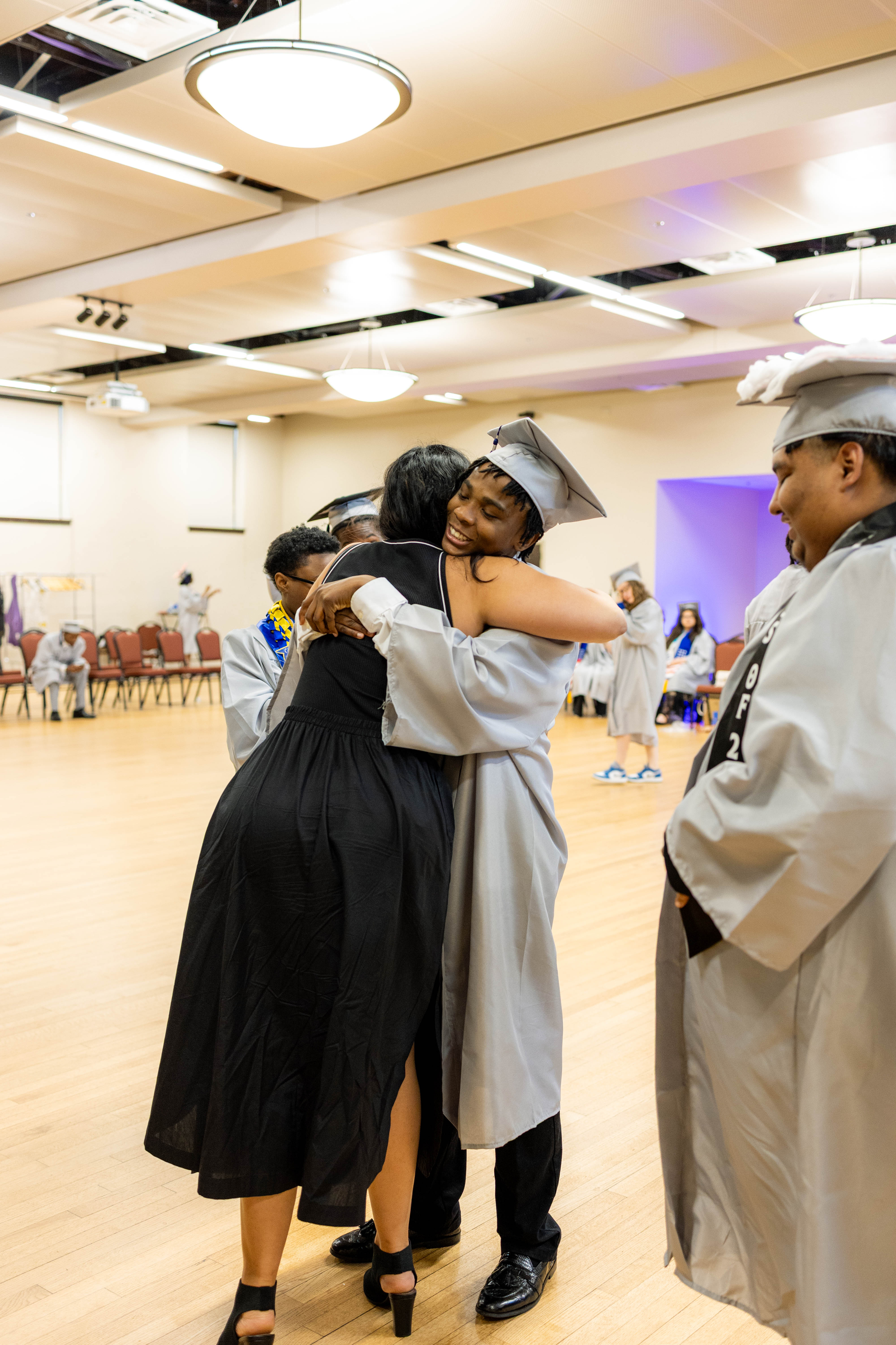 Group of graduates in caps and gowns hugging each other in a large room with a hardwood floor, chairs, and bright ceiling lights.