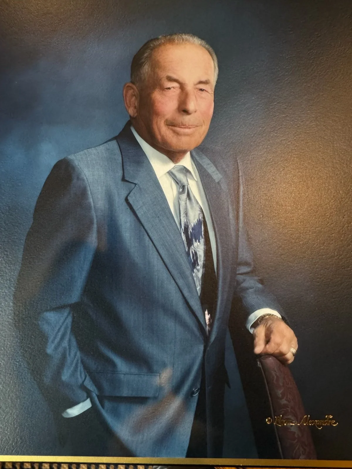 A portrait of an elderly man in a gray suit and patterned tie, standing against a dark background.