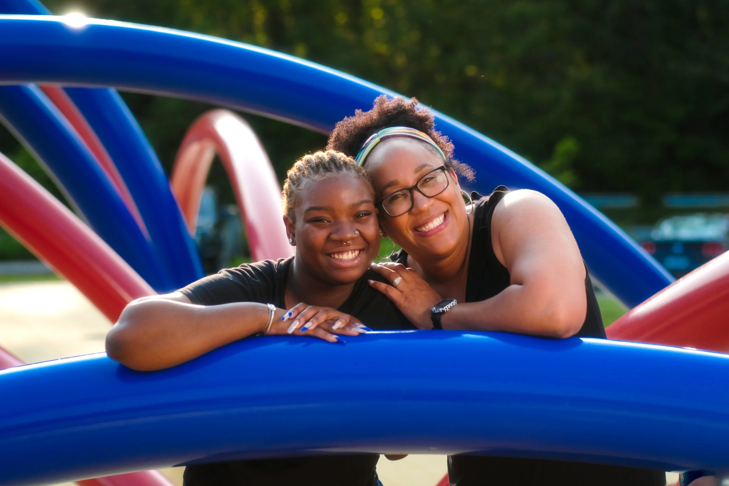 Two women smiling and leaning on a blue playground structure in an outdoor park with trees in the background.