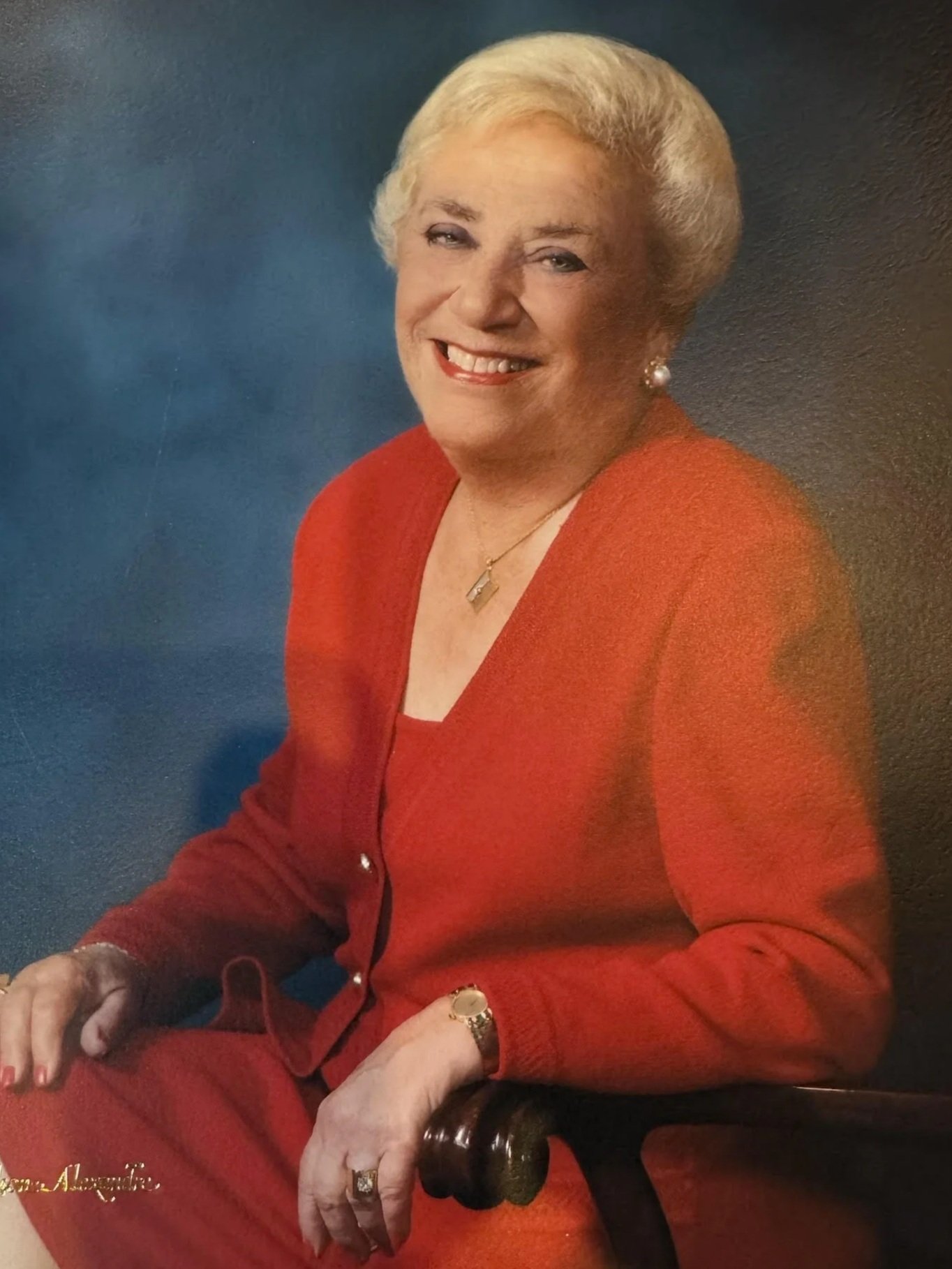 A woman with short blonde hair, wearing a red blazer, pearl earrings, and a gold necklace, smiling at the camera.