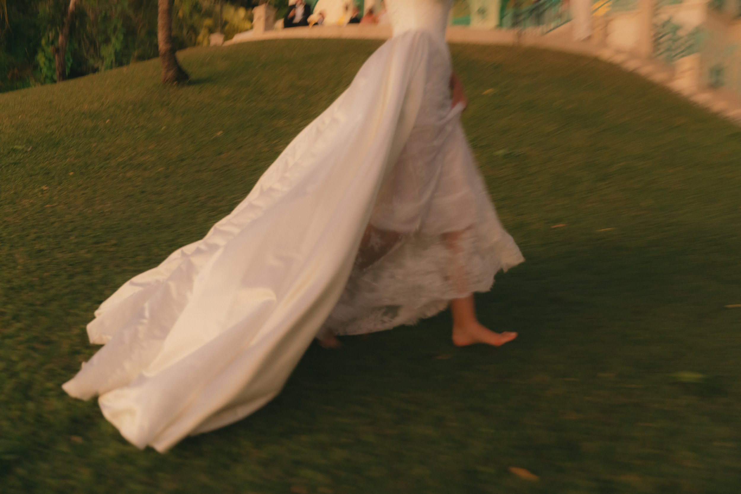 A child walking barefoot on grass, wearing a lace dress and holding a white fabric or veil, at night or in low light.