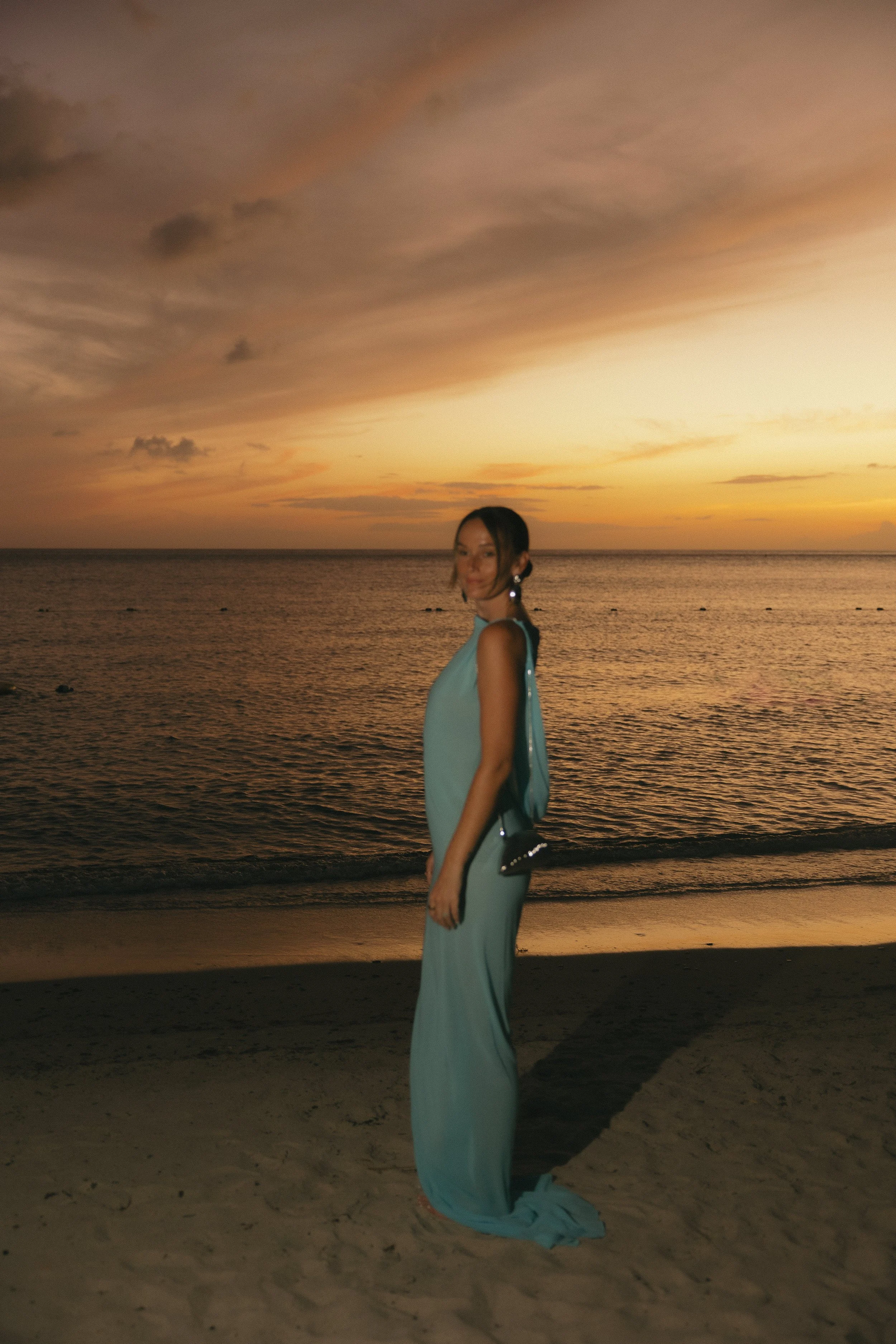 A woman in a long teal dress standing on a beach at sunset, holding a small purse, with the ocean and colorful sky in the background.