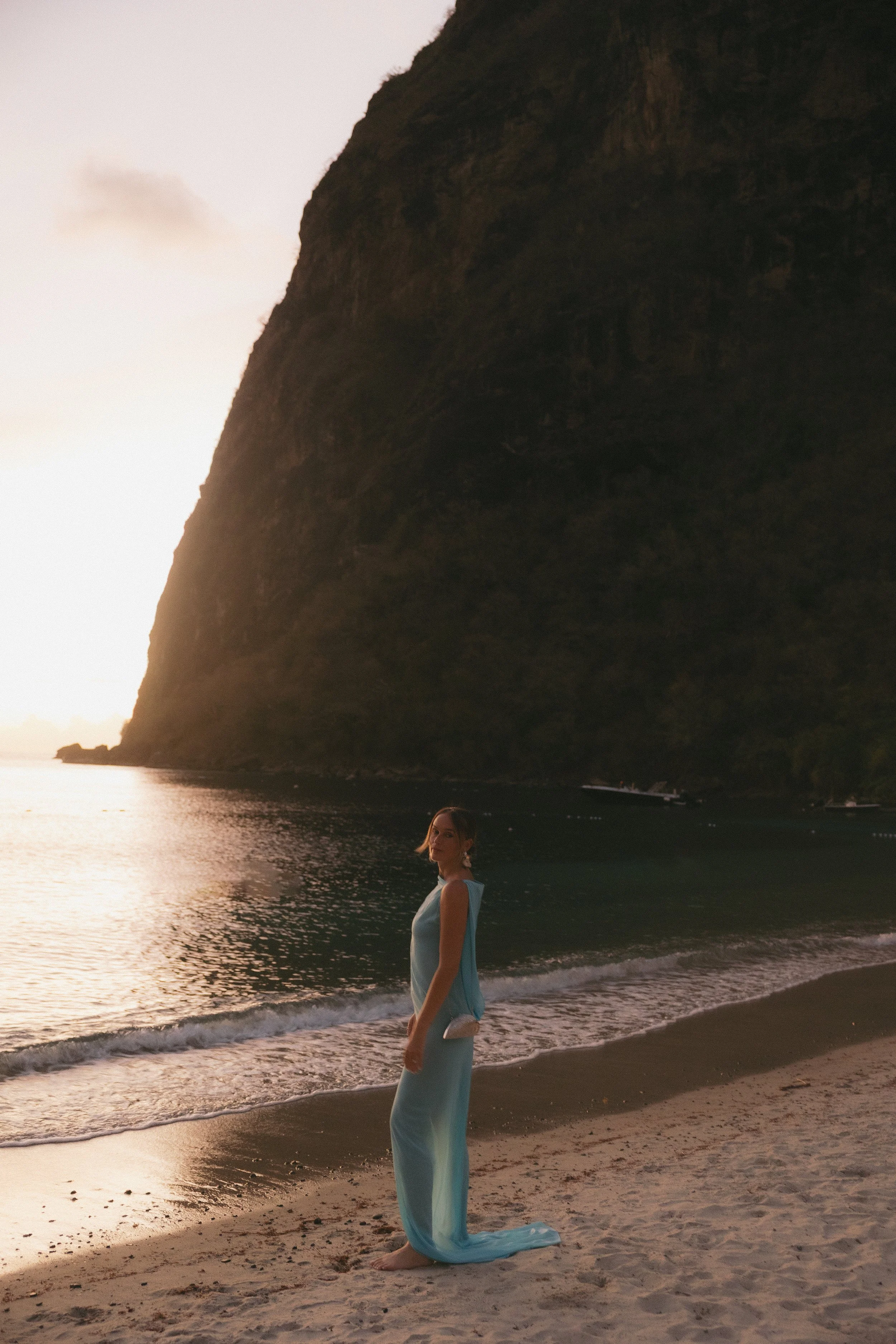 A woman in a long blue dress standing on a beach at sunset, with a large rocky cliff in the background and calm water reflecting the sky.
