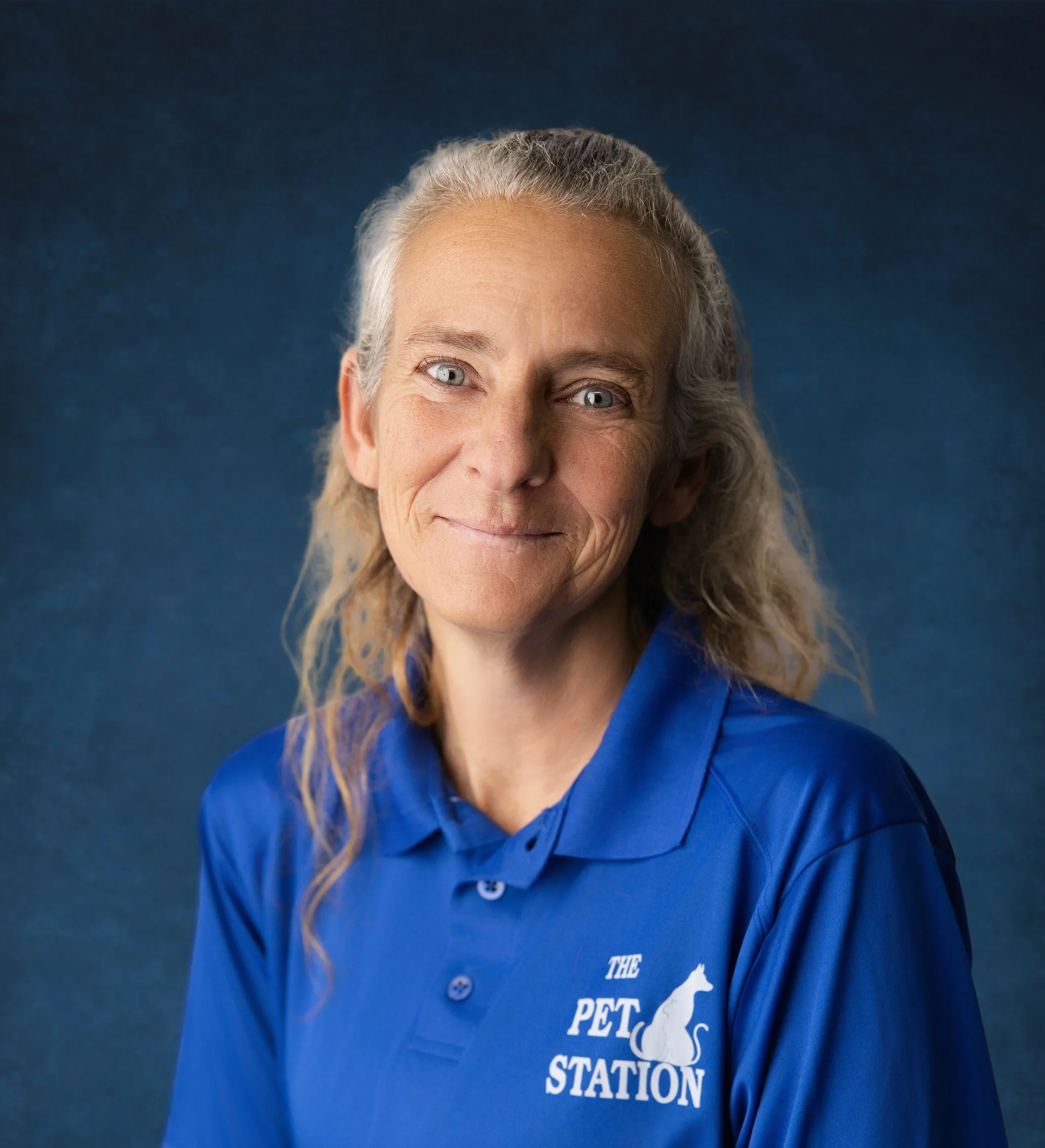 Close-up of a smiling woman with gray hair, blue eyes, and fair skin, wearing a blue shirt with white text that reads 'The Pet...' against a dark blue background.