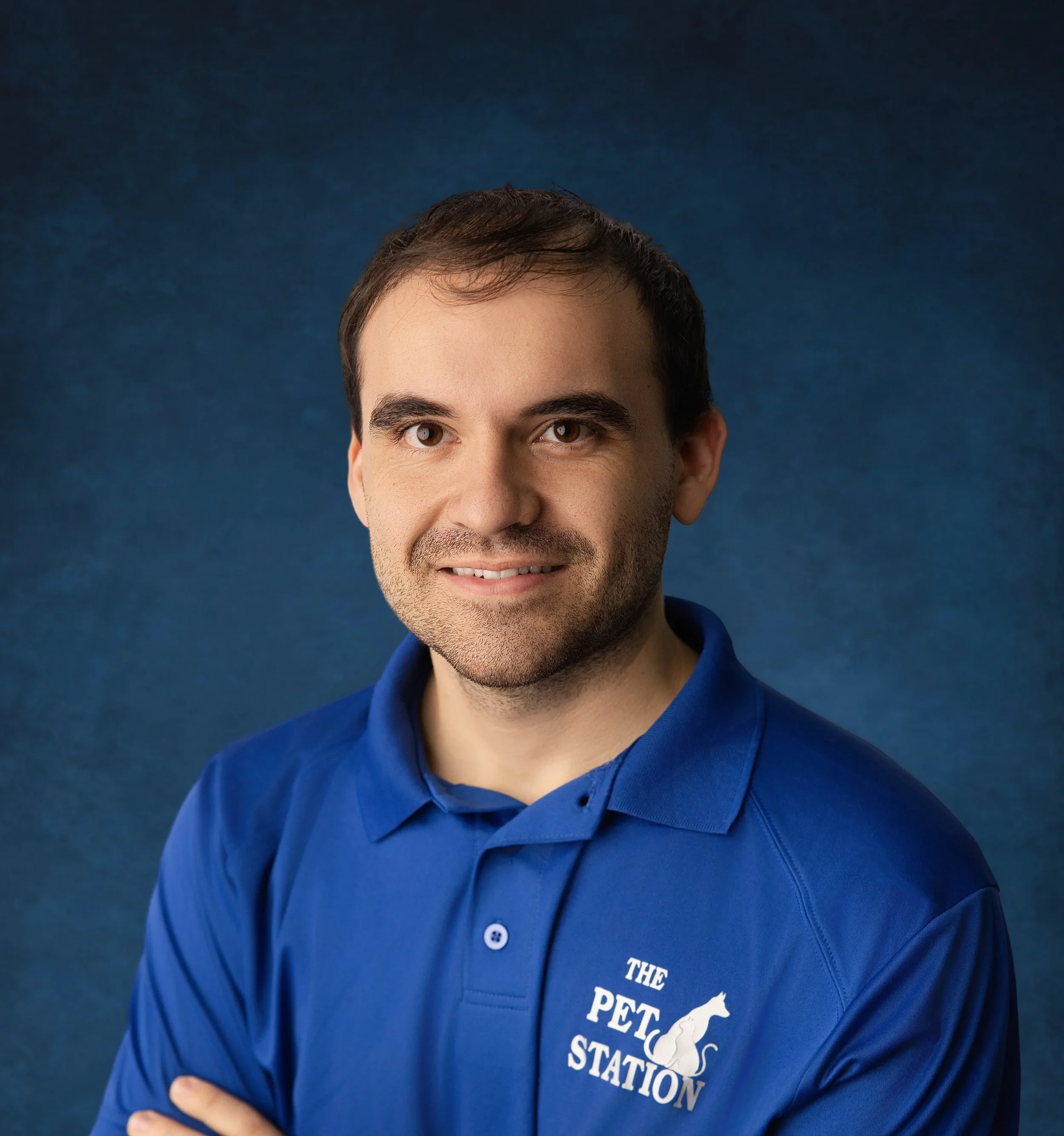 Close-up of a man wearing a navy blue shirt with a logo that reads 'The Pet Station' and a silhouette of a dog and cat, posing with arms crossed against a blue background.