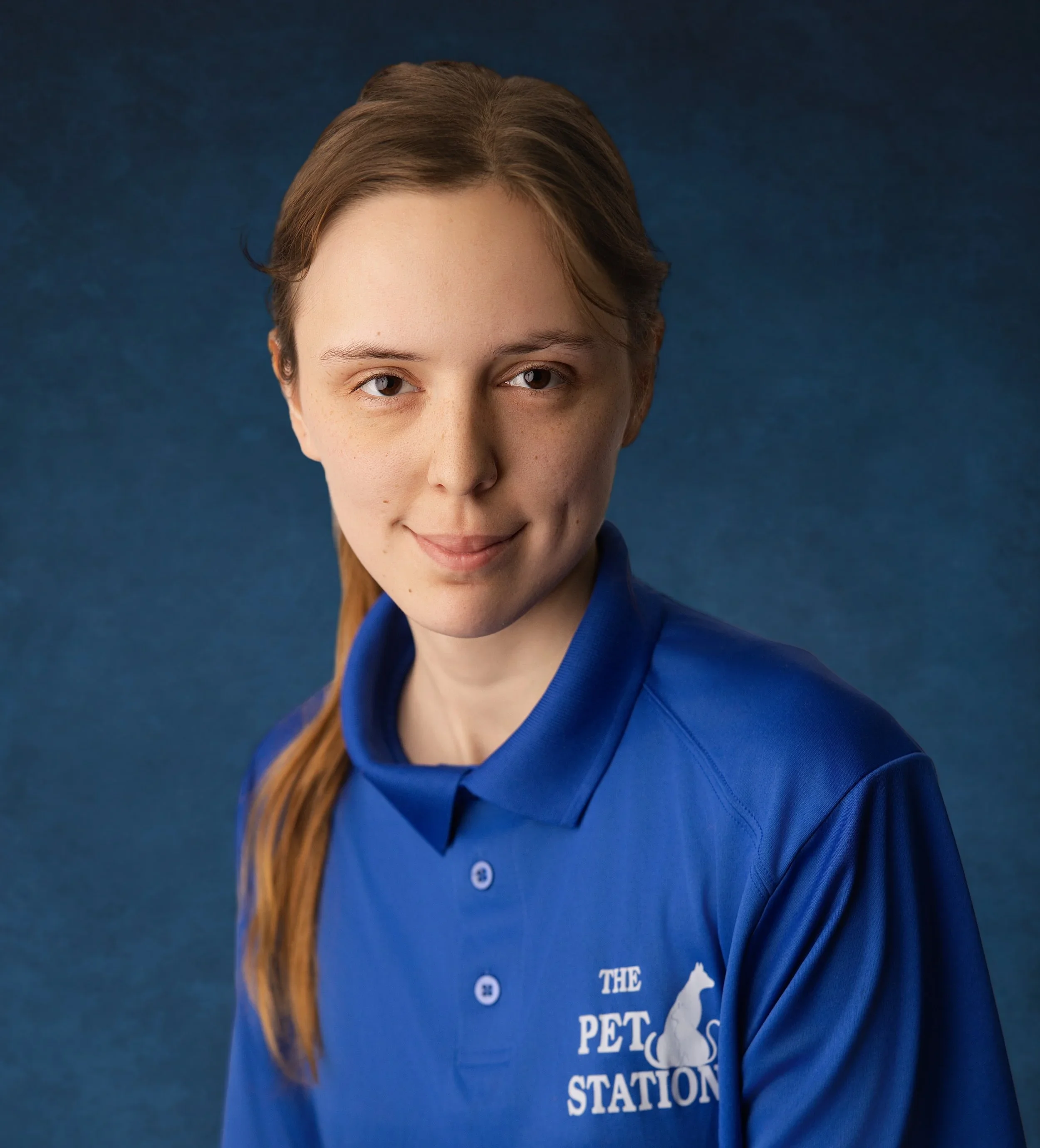 Close-up of a woman's face, with brown hair tied back, wearing a blue jacket with white text and graphics, against a dark blue background.