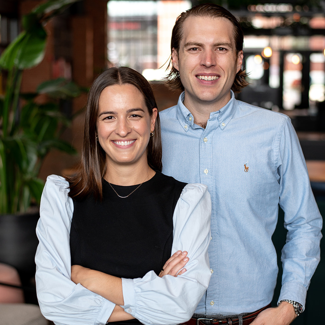 A smiling woman with shoulder-length dark brown hair, wearing a black dress with white puffed sleeves and a silver necklace, standing next to a smiling man with shoulder-length light brown hair, wearing a light blue button-up shirt with a small emblem, in a cozy, modern indoor setting with plants and warm lighting.