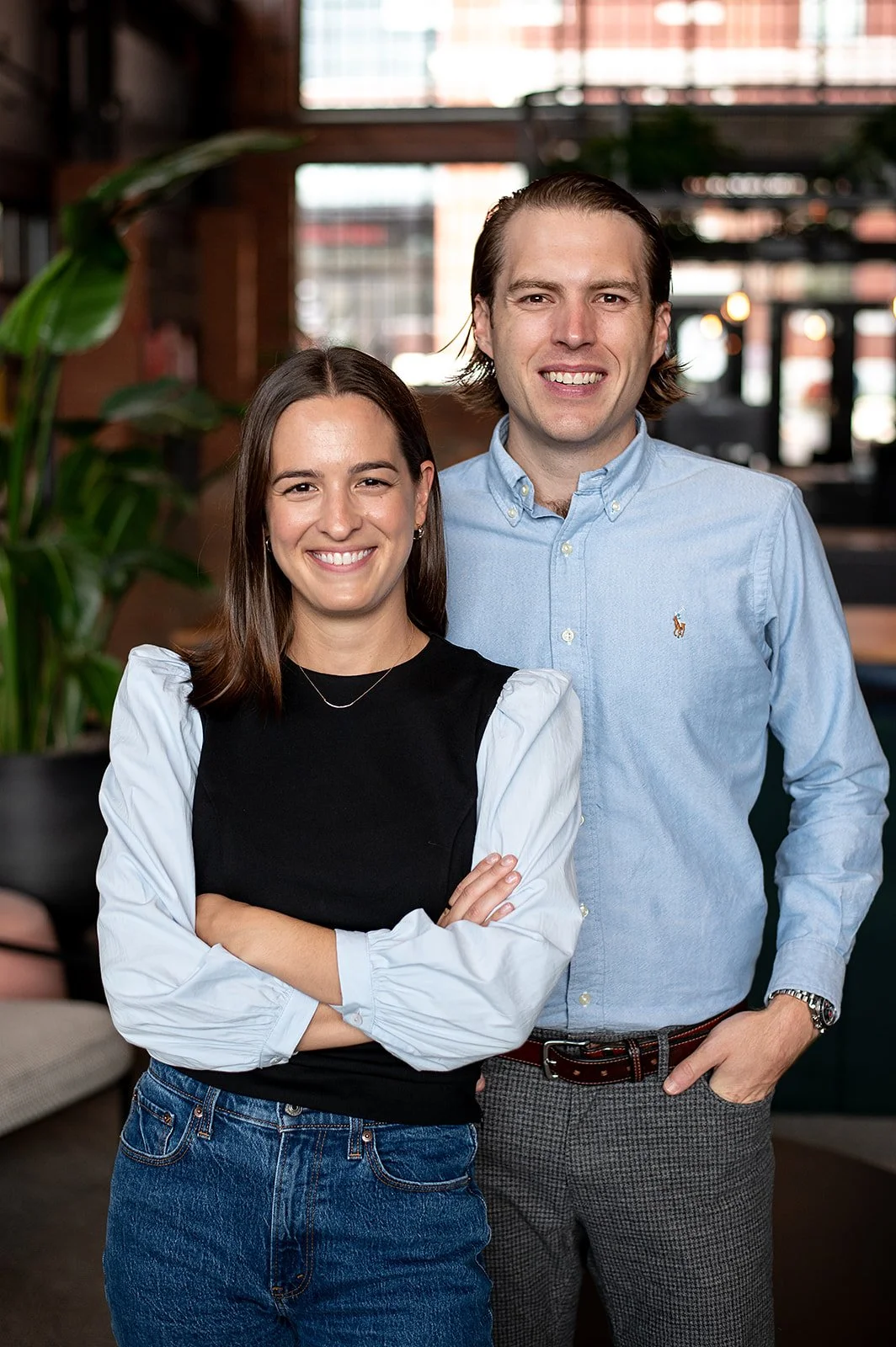 A woman with shoulder-length brown hair, wearing a black top with white sleeves and blue jeans, stands with her arms crossed, smiling. A man with medium-length brown hair, wearing a light blue button-down shirt and gray pants, stands behind her with a smile. They are indoors in a well-lit space with large windows and green plants in the background.