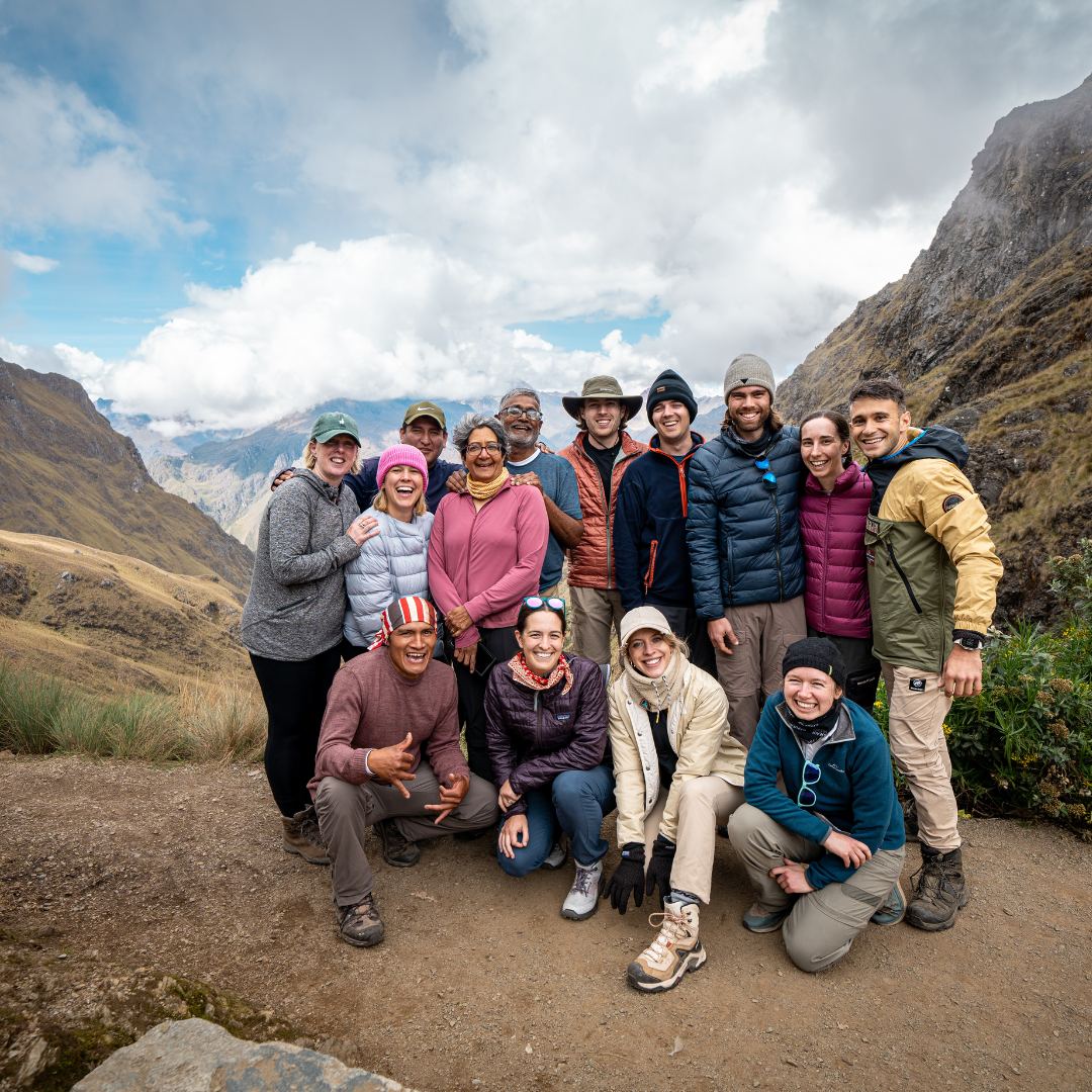 Group of hikers smiling and posing for a photo in a mountainous landscape with clouds and blue sky in the background.