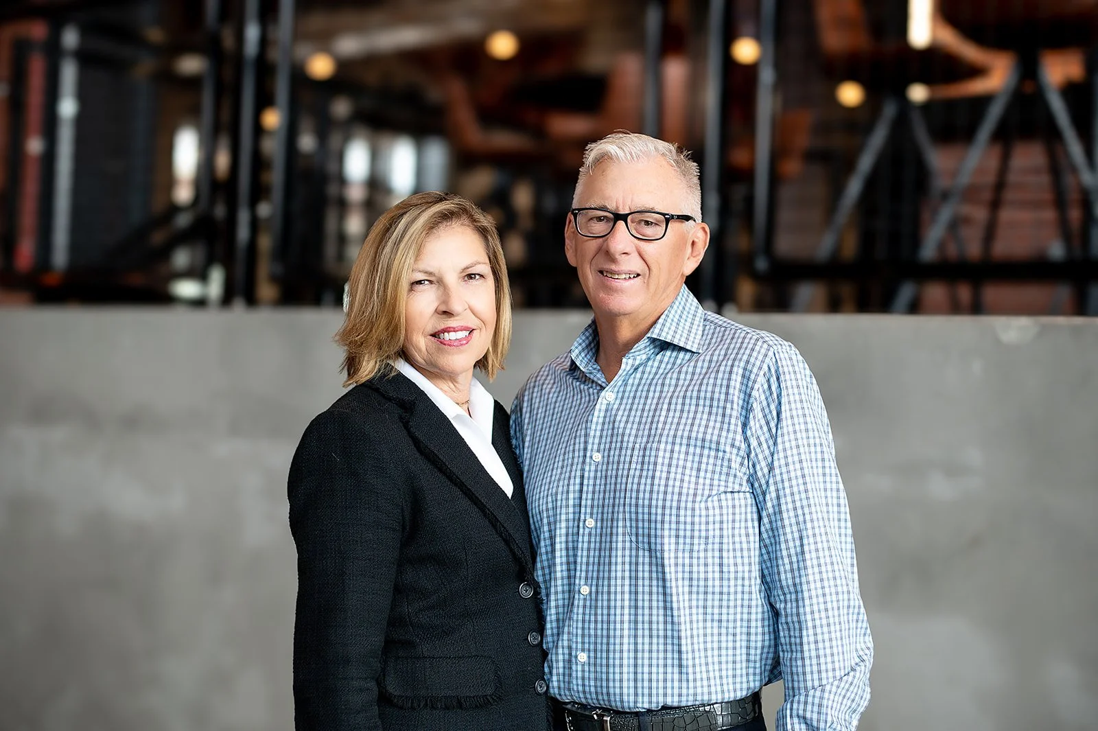 A photo of a middle-aged woman with blonde hair wearing a black blazer and a middle-aged man with gray hair, glasses, and a checked shirt standing together indoors.
