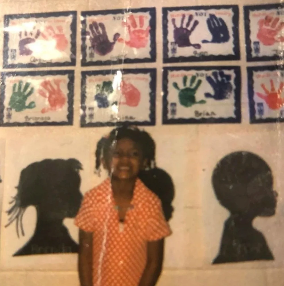 A young, proud Dr. Brenda smiling in front of a wall of class art