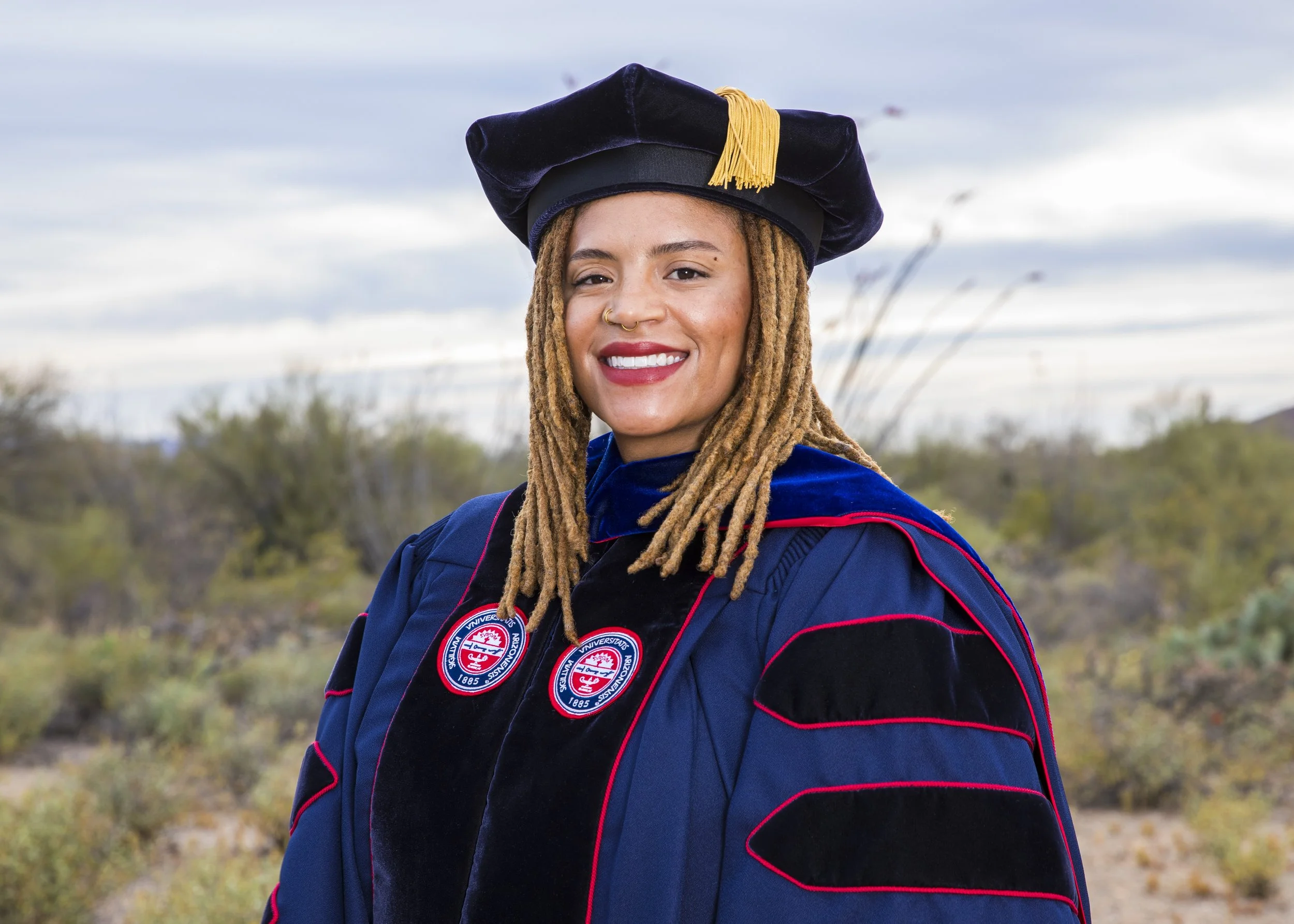 A young woman with dreadlocks smiling outdoors wearing a navy blue academic gown with patches and a matching cap with a yellow tassel.