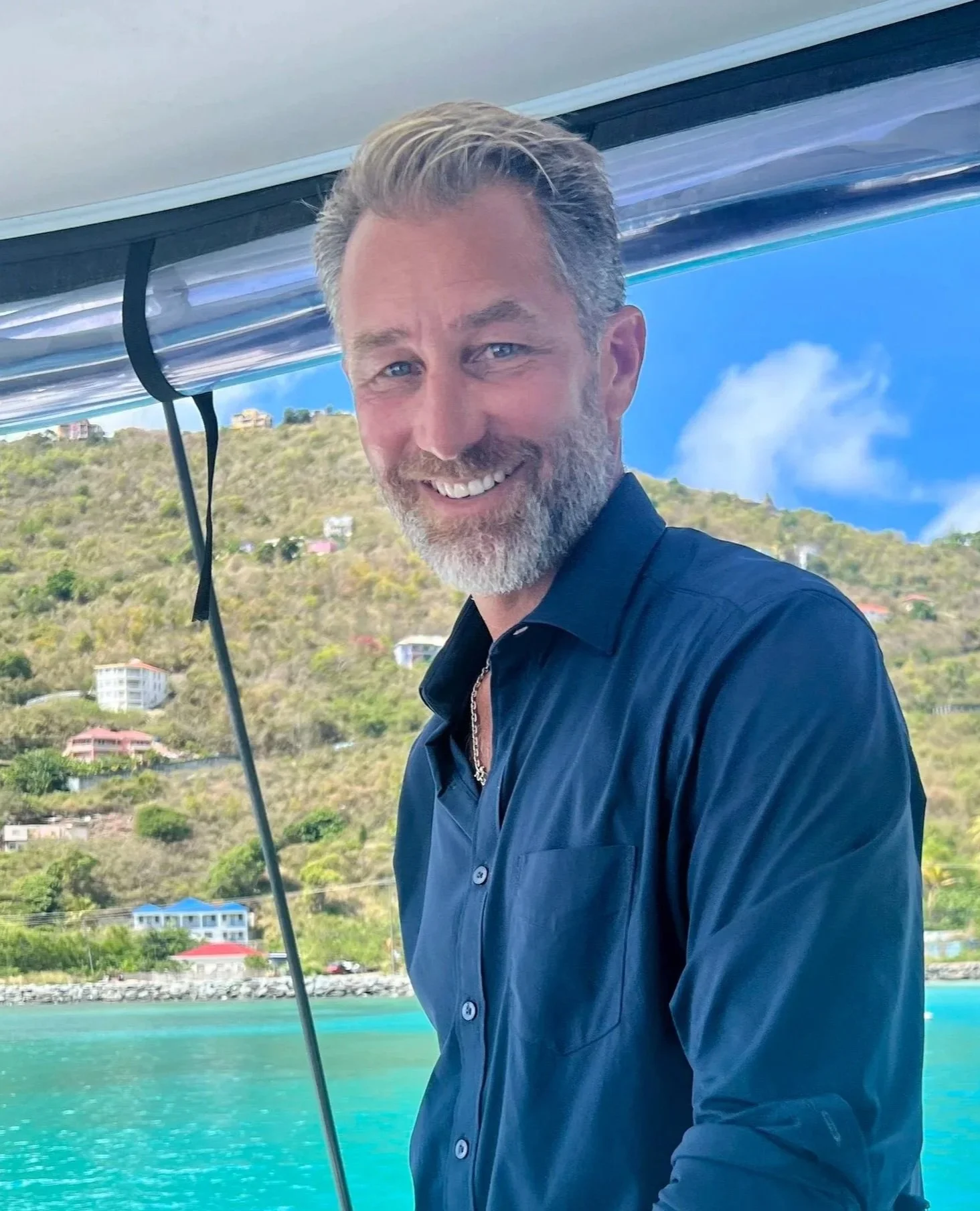 Smiling man with gray beard and blue eyes in a navy shirt on a boat, with hillside and houses in the background under a blue sky.