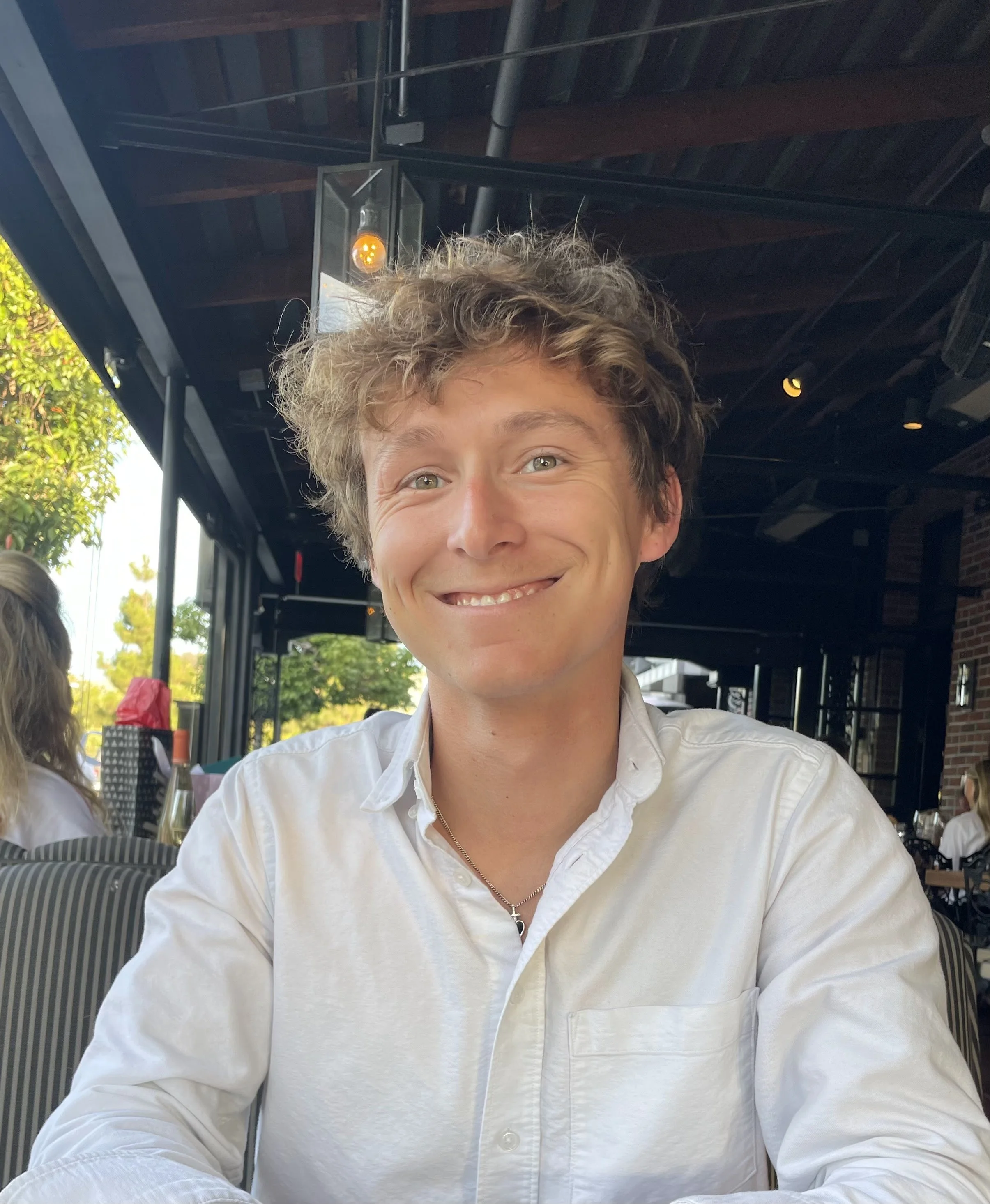 A young man with curly light brown hair smiling with bright eyes, wearing a white button-up shirt, sitting at an outdoor cafe with green trees and a building in the background, under a black metal roof.